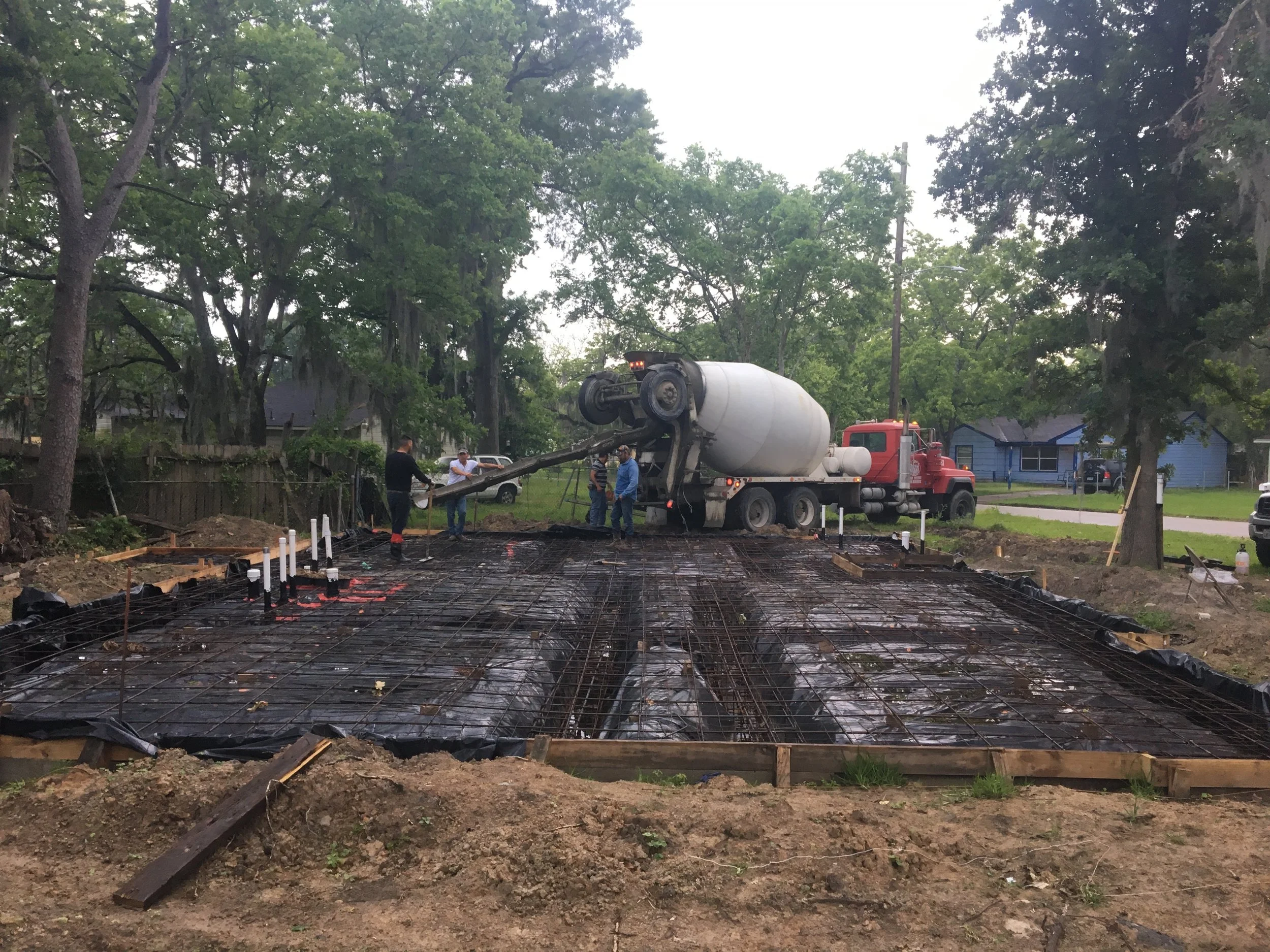 Construction workers and a cement mixer truck working on laying a concrete foundation in a residential neighborhood surrounded by trees.