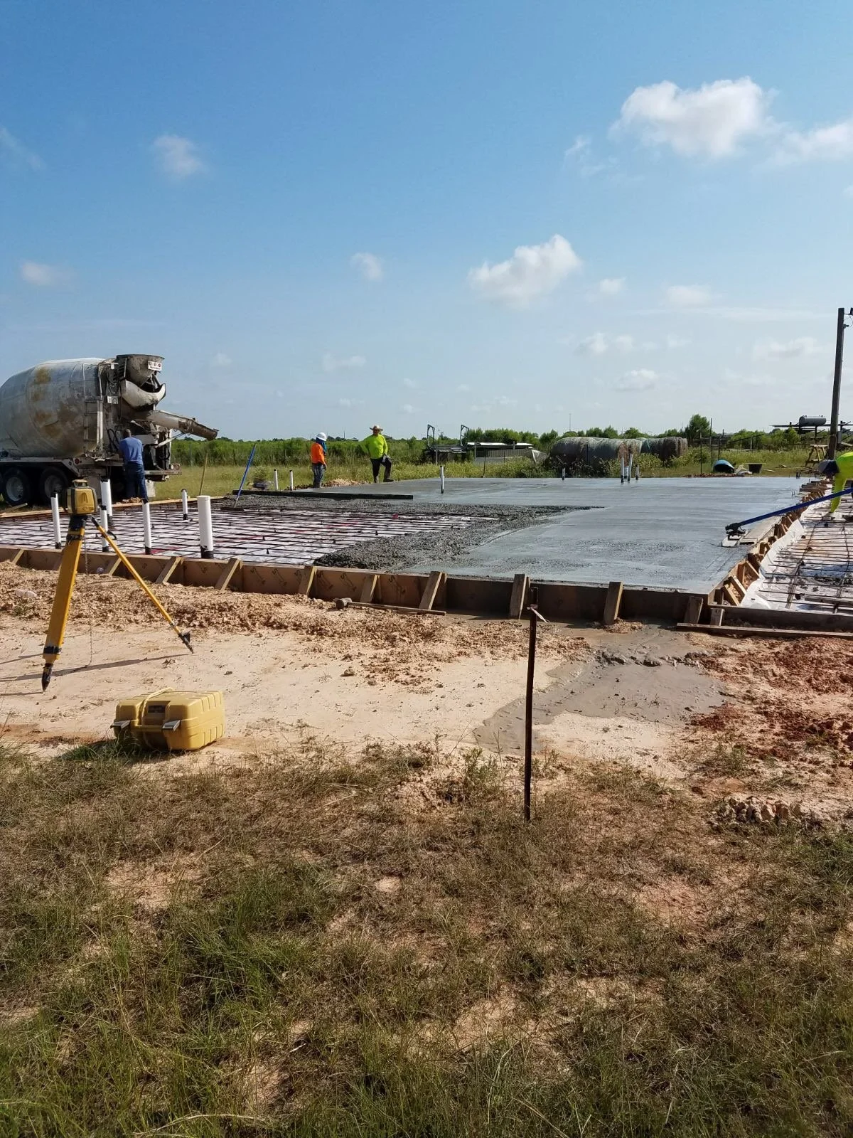 Workers pour concrete on a construction site with tools and equipment, including a cement truck and measuring instruments, in a rural area with blue sky and green vegetation.