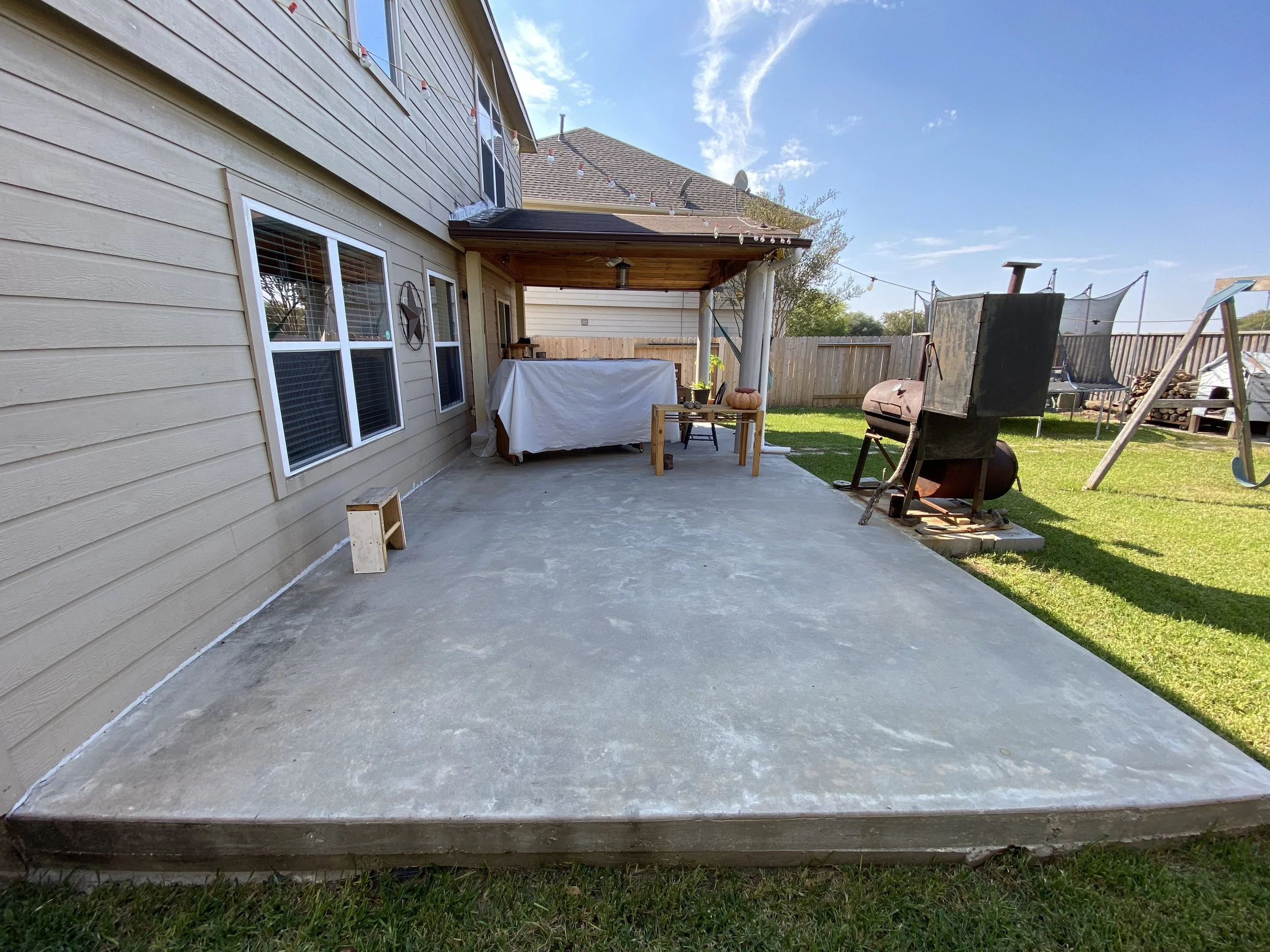 Backyard patio area with a concrete floor, outdoor table with a white cover, pumpkin, and a small wooden bench. There is a nearby grill, a swing set, trampoline, and wood pile in the yard, with a wooden fence surrounding the yard and a house with beige siding.