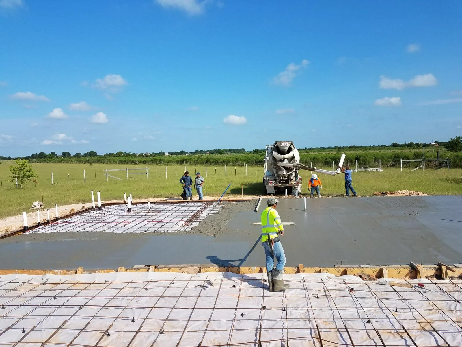 Construction workers pouring concrete on a building foundation in a rural area, with a concrete mixer truck and blue sky in the background.