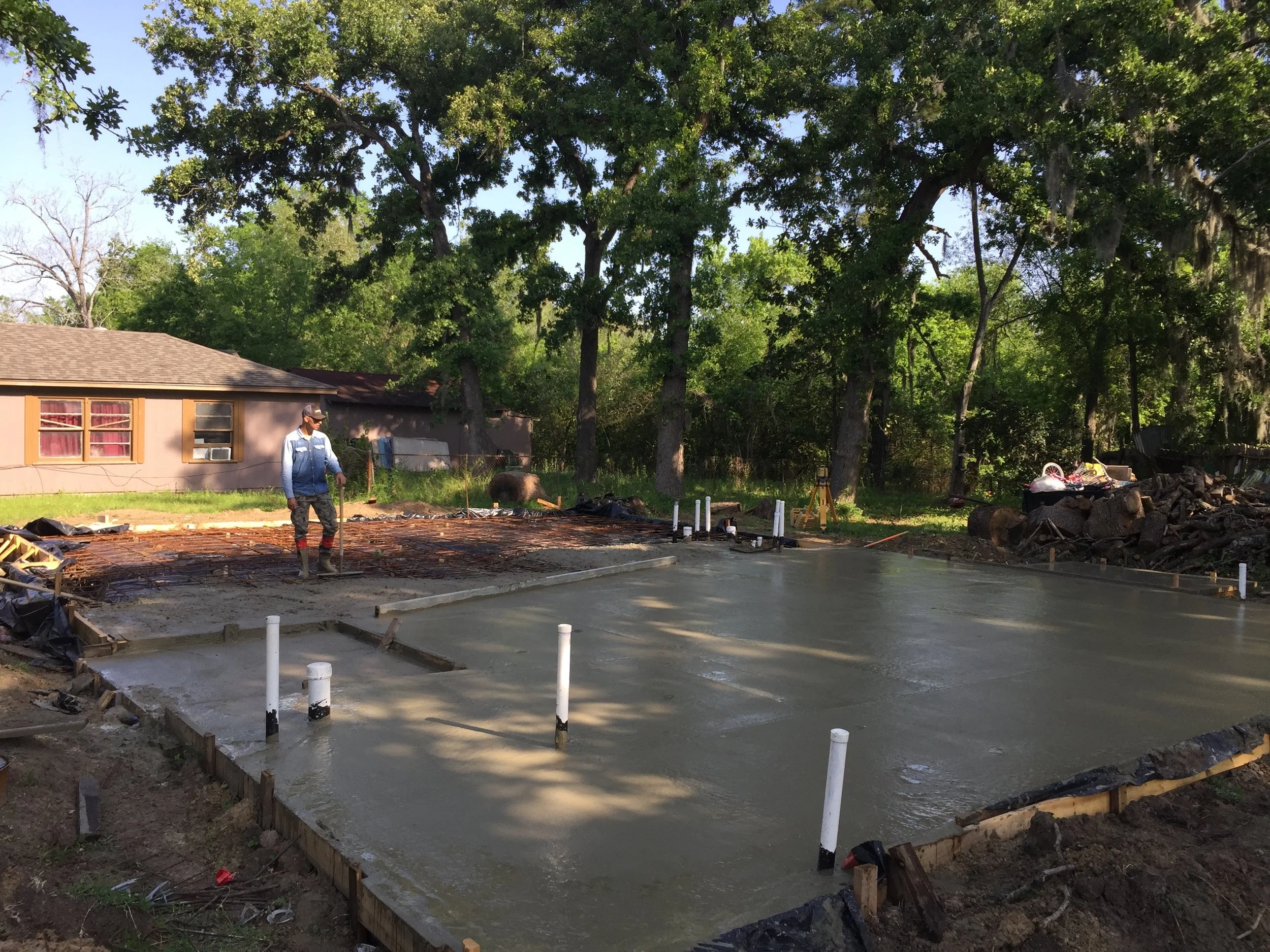 A man working on a small concrete slab in a backyard as part of construction or landscaping work, with a house and tall trees in the background.
