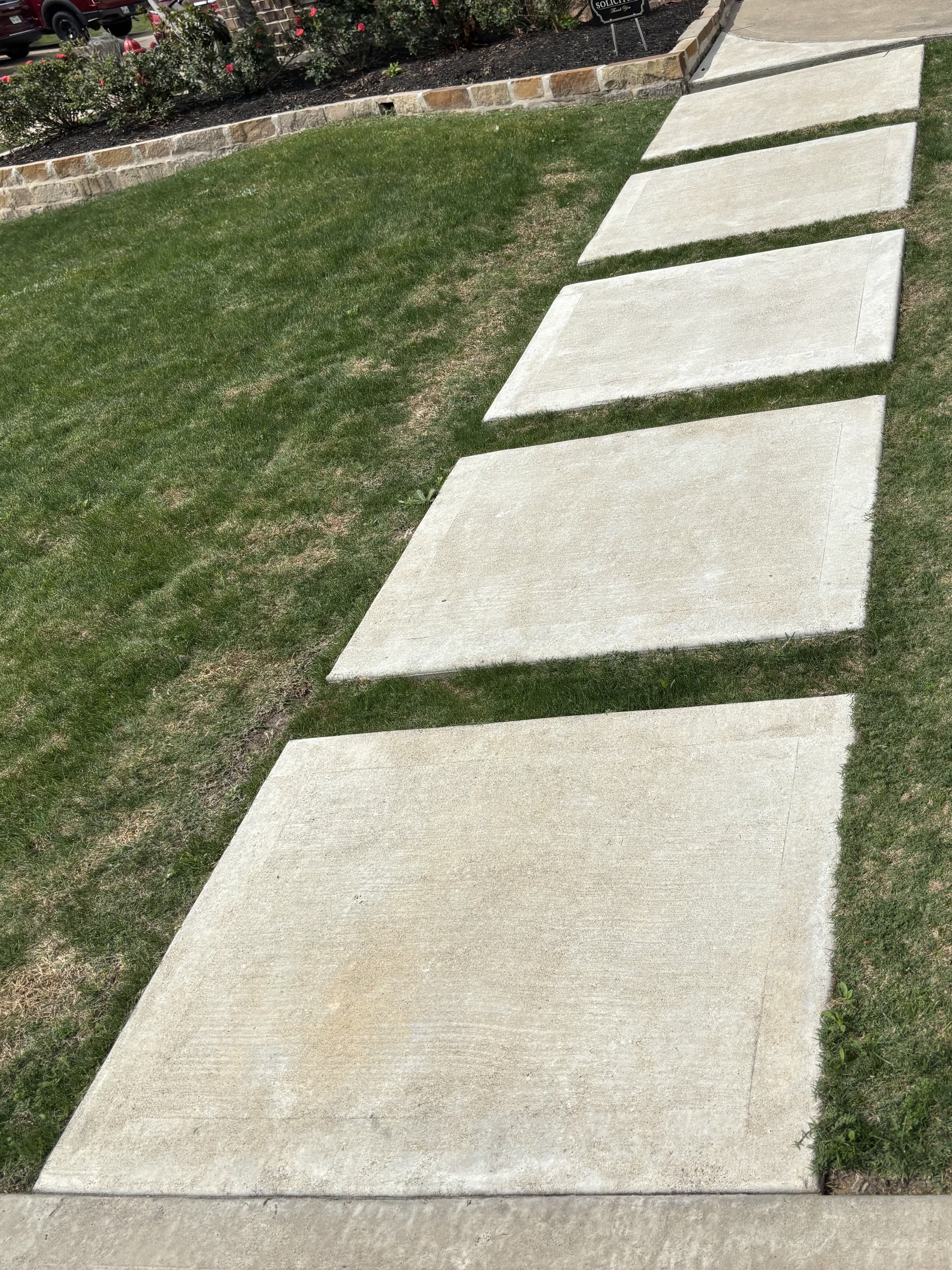 Concrete sidewalk with large rectangular slabs on a grassy lawn, bordered by a low brick wall and flowering bushes in the background.