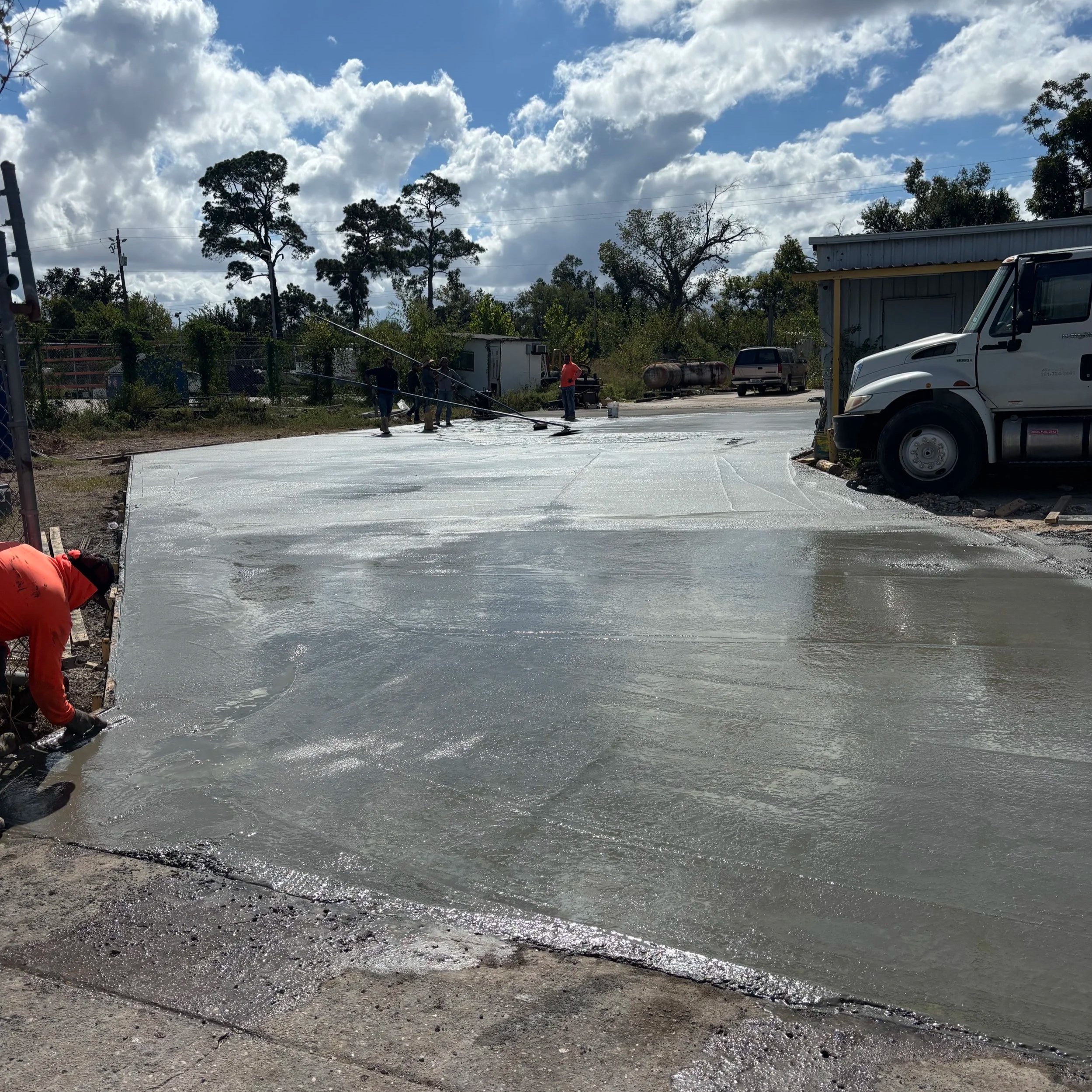 Workers applying a fresh layer of concrete on a sidewalk outdoors, with a blue sky and scattered clouds overhead, and trees and utility poles in the background.