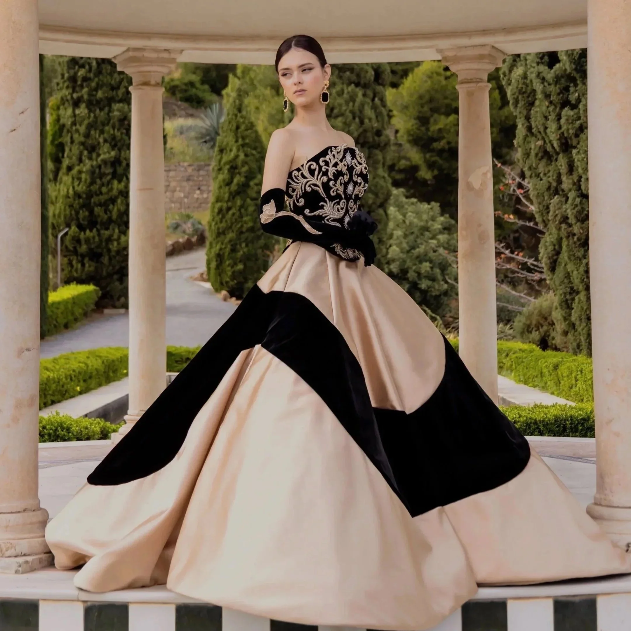 A woman dressed in an ornate black and white gown, standing under a classical stone portico with columns, with a green, landscaped background.