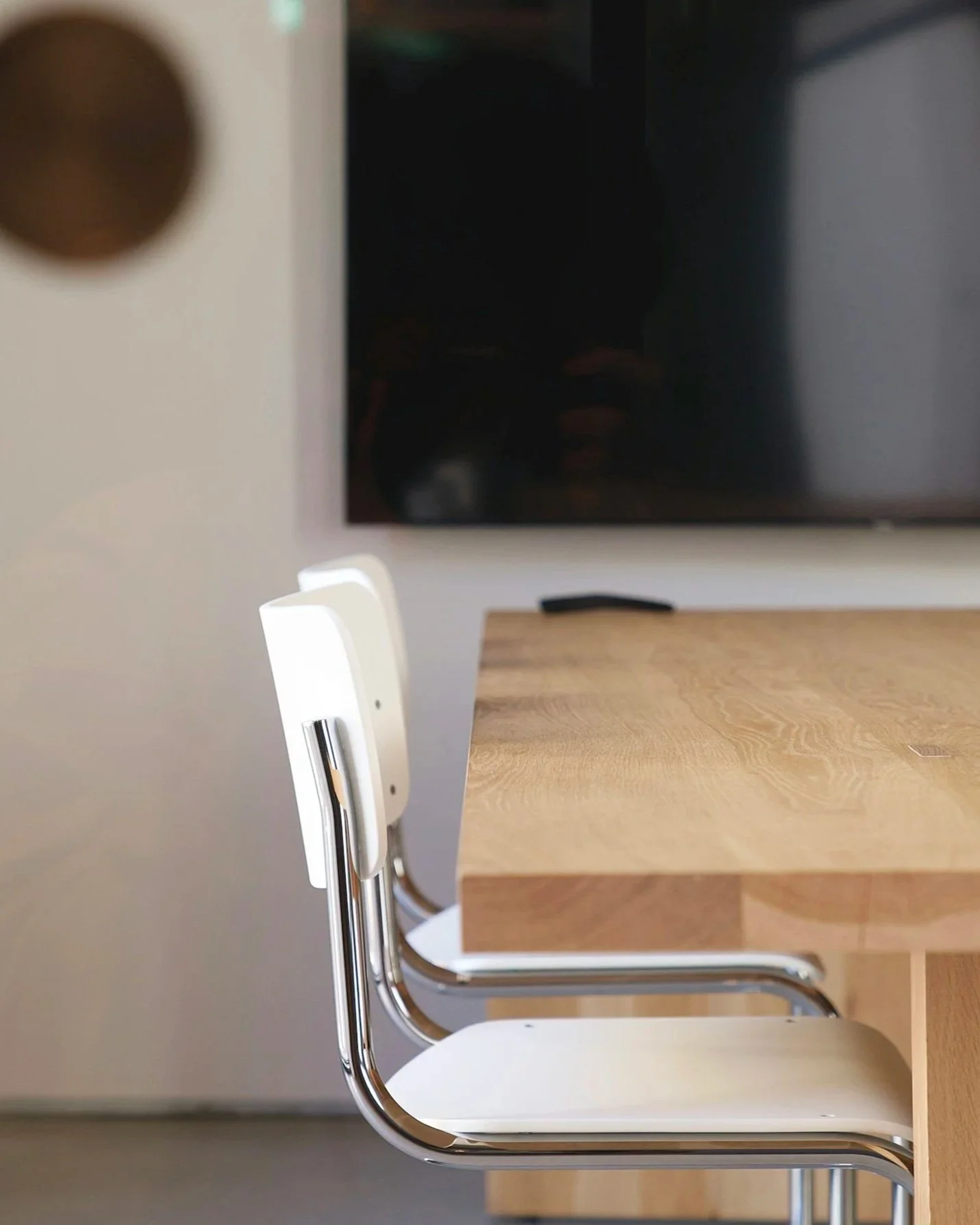 The image shows a close-up view of a white and metallic chair next to a light wooden table, with a black, flat-screen monitor in the background.