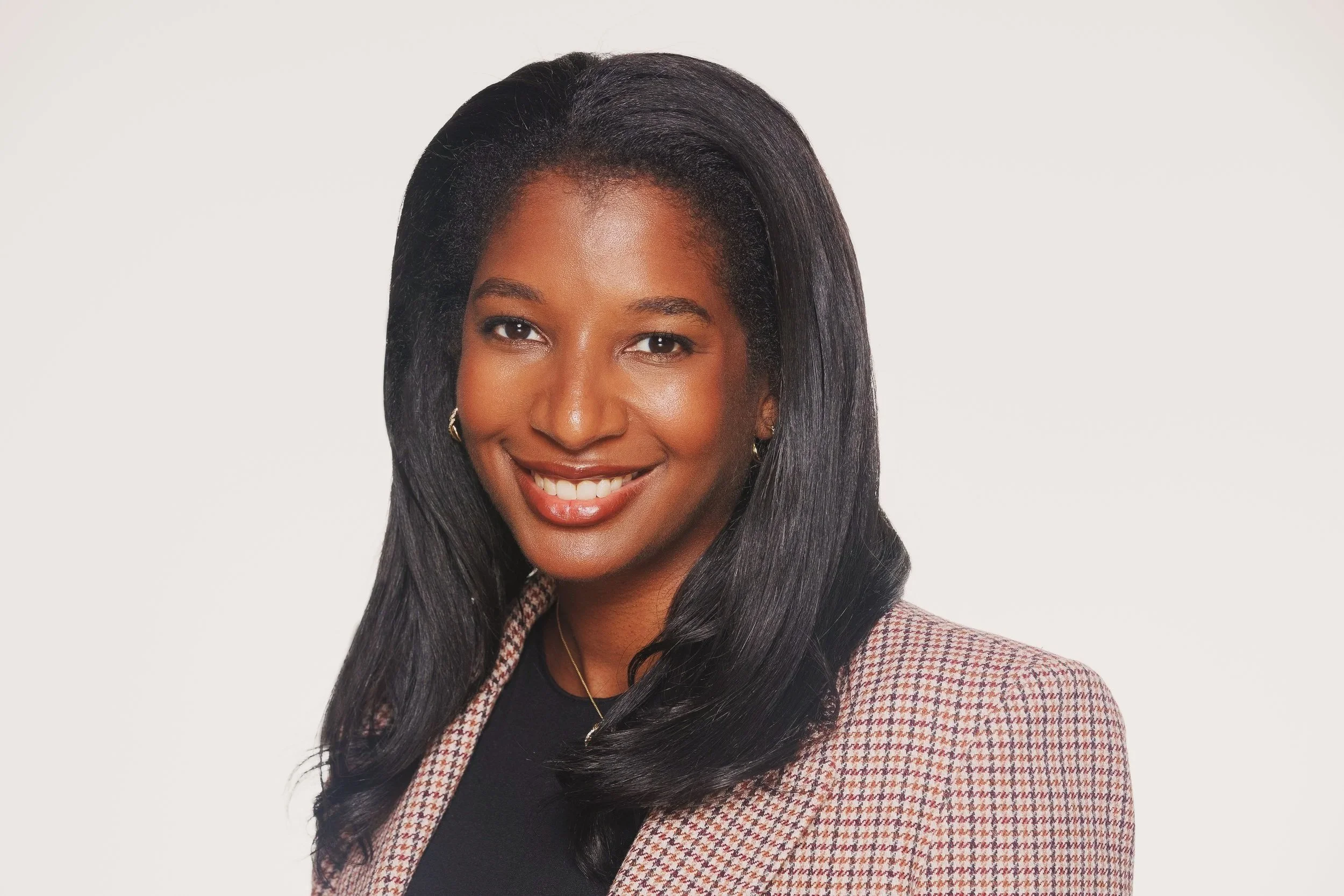 A woman with long black hair smiling, wearing a plaid blazer, gold earrings, and a black top, against a plain light background.