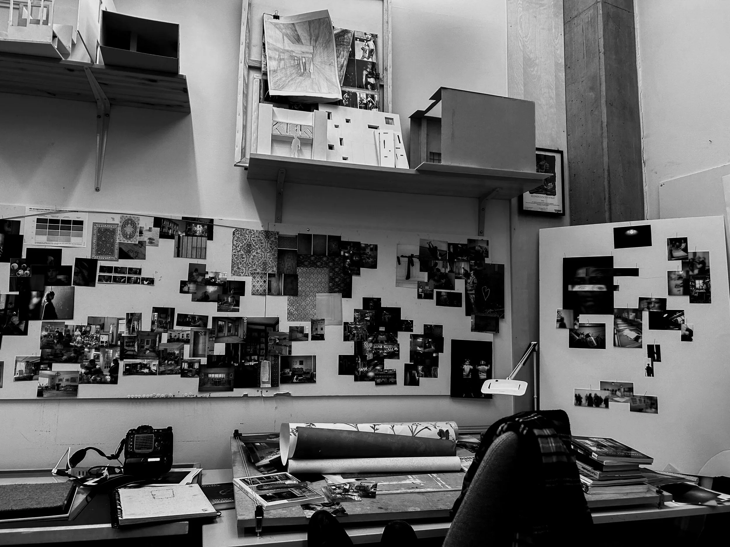 A workspace with a desk, a chair, and various items, including a camera, papers, and a roll of paper. The wall behind the desk is decorated with numerous photographs, posters, and artwork, and there are shelves with architectural models and framed pictures. The setting appears to be an artist's or architect's studio.