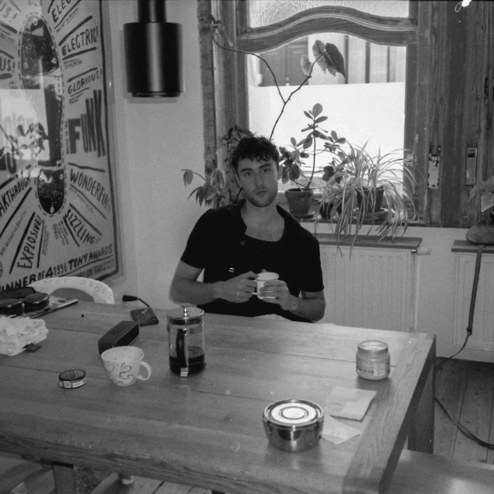 A young man with dark curly hair sitting at a wooden table holding a cup. The table has various items including a glass, a small bowl, a pack of cigarettes, and some papers. Behind him, there are potted plants on a window sill and a large poster on the wall. The scene is indoors with a mix of modern and rustic decor.