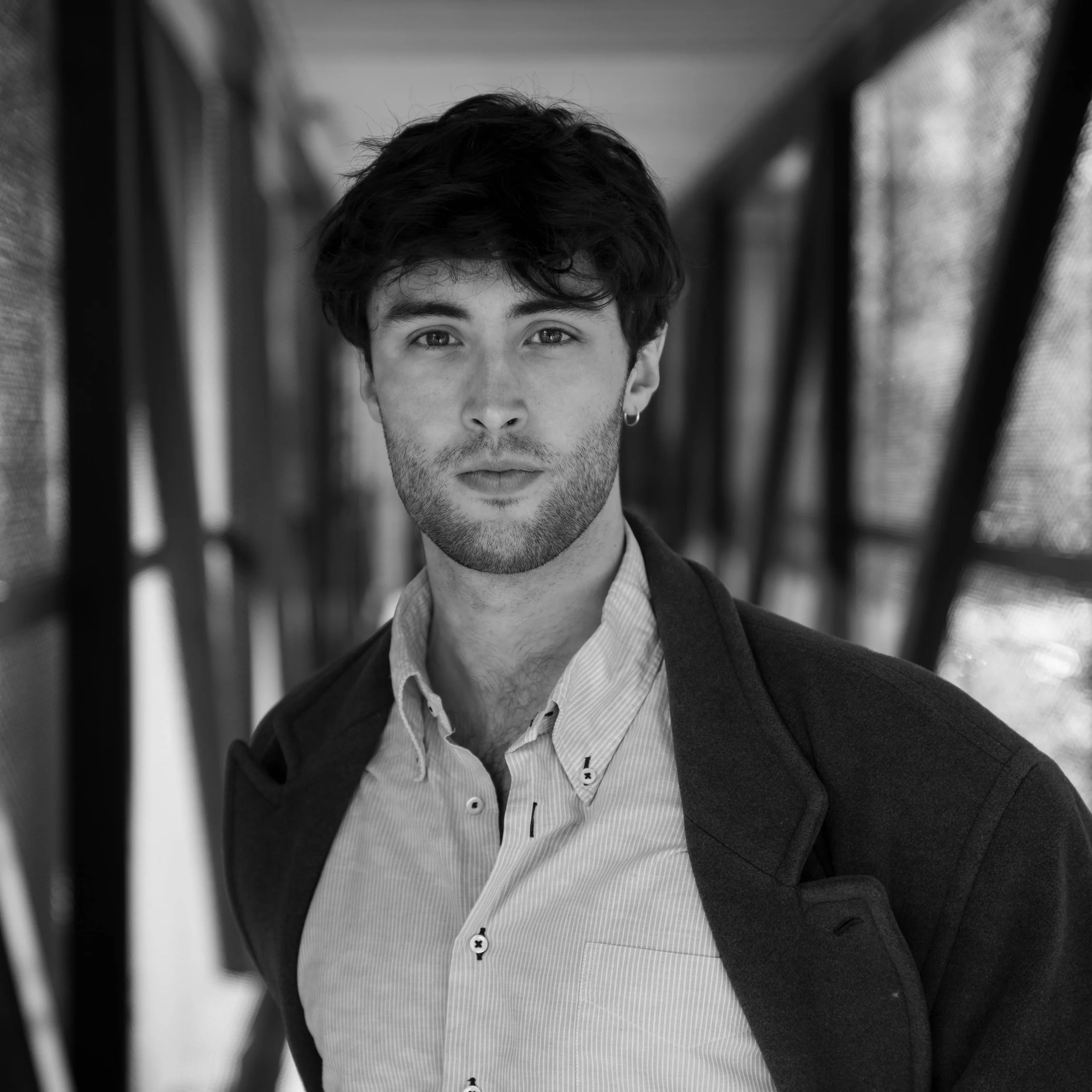 Black and white close-up of a young man with wavy dark hair, wearing a white collared shirt and a dark blazer, standing outdoors between two structures with blurred background.