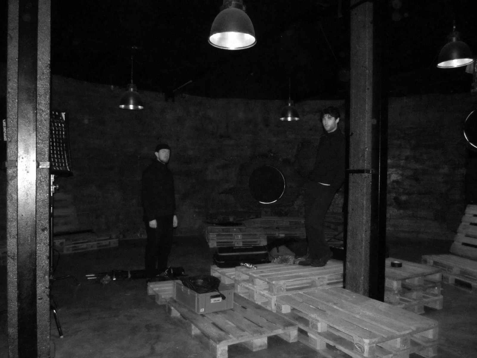 Two men in a dark industrial space with wooden pallets, some musical equipment, and hanging pendant lights.