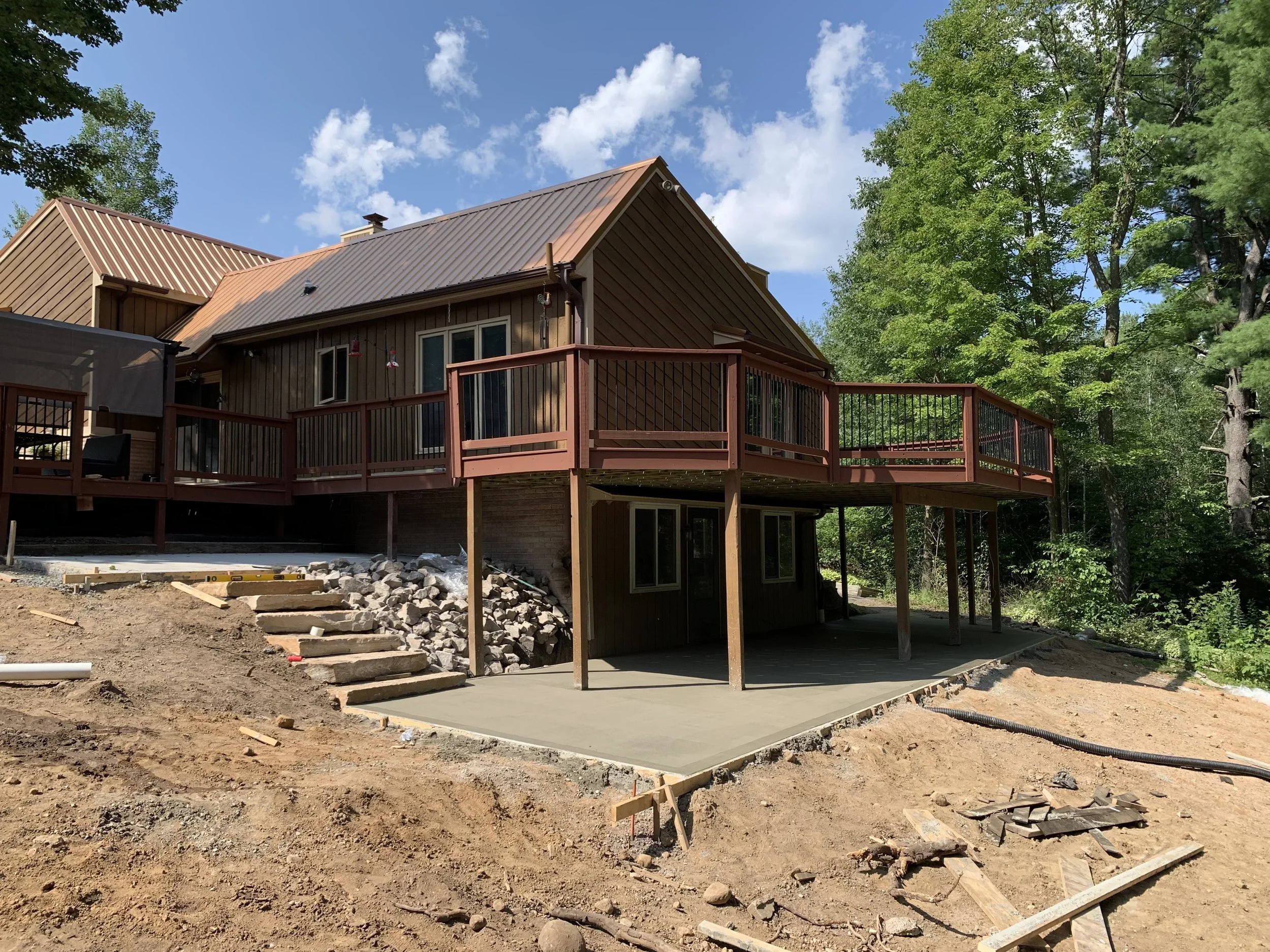 The image shows the backyard of a house under construction. It features a newly poured concrete slab, some stone steps, and a wooden deck with black metal railings attached to the house. The house has a brown metal roof and dark brown siding. The surroundings include dirt and construction debris, along with green trees and a partly cloudy blue sky.