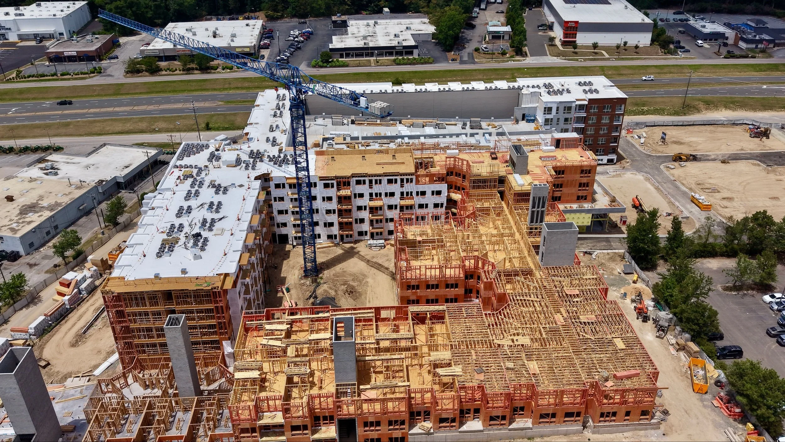 An aerial view of a construction site with multiple buildings in different stages of construction, a tall blue crane, and nearby roads and parking lots.