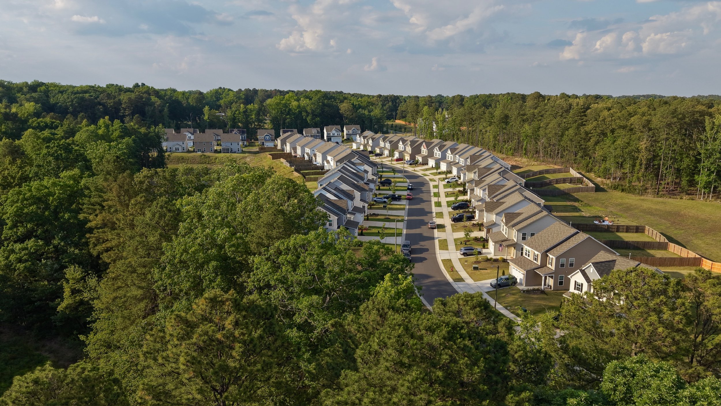 An aerial view of a suburban neighborhood with rows of houses, surrounded by green trees and forested areas, under a partly cloudy sky.