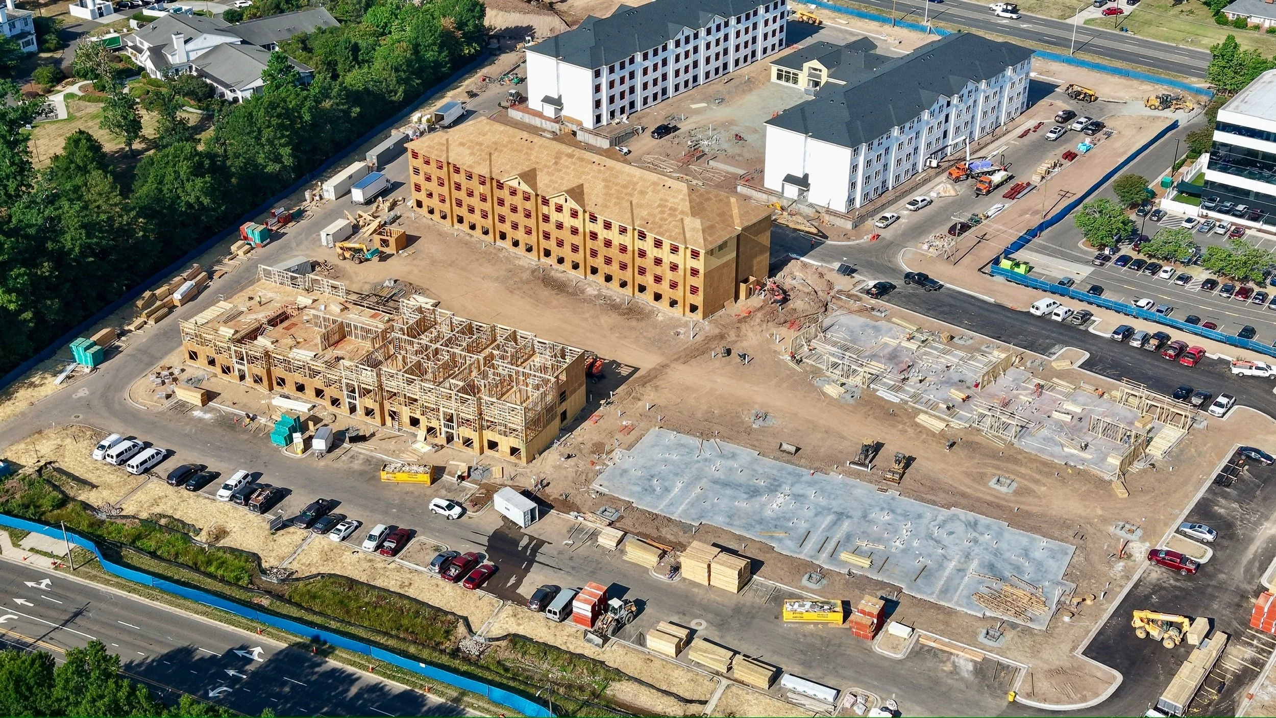 An aerial view of a construction site with multiple buildings in various stages of completion, surrounded by parking lots and green trees.