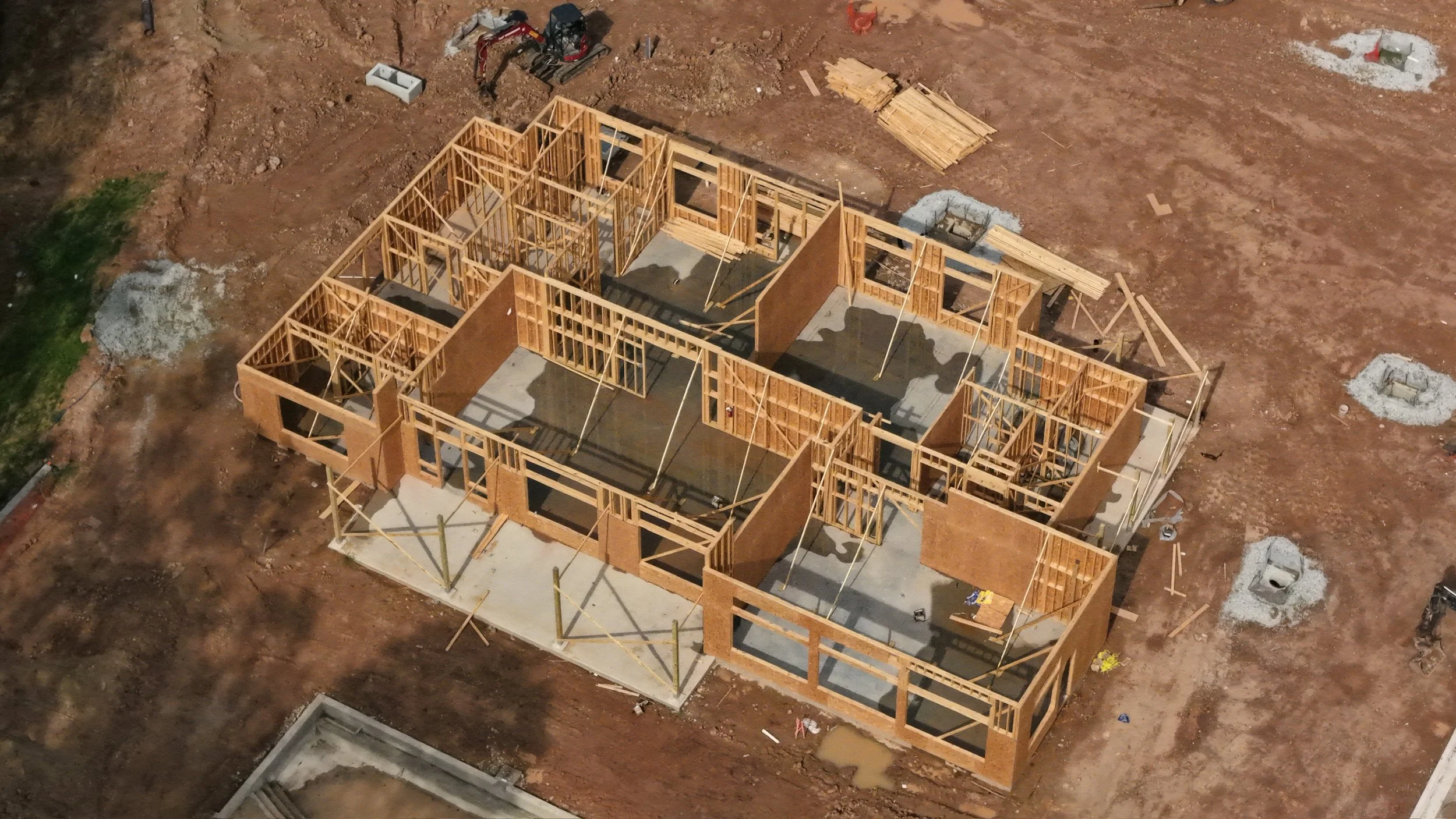 Construction site showing wooden framework of a house being built, with surrounding dirt and construction materials.