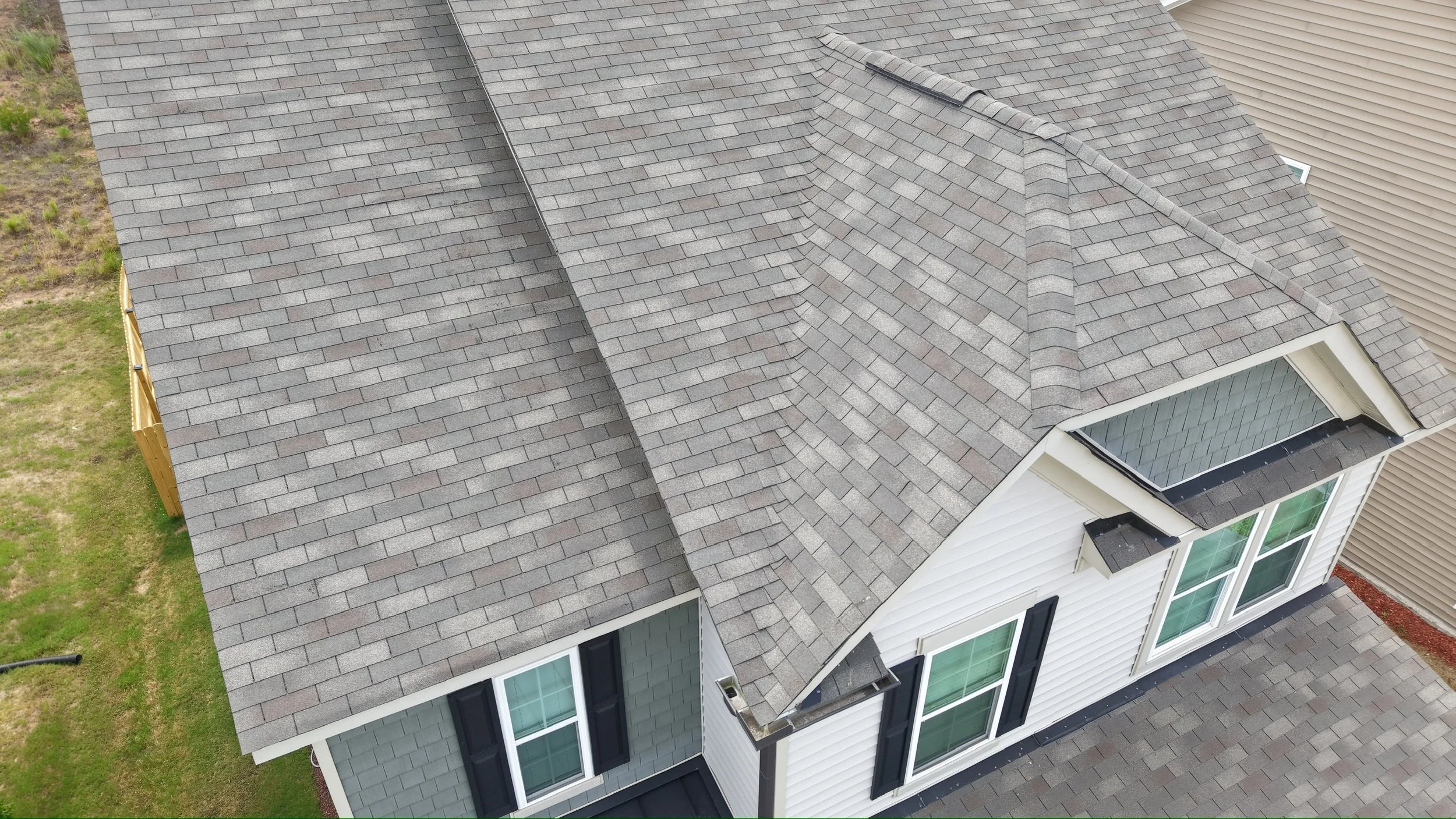 Aerial view of a house roof with gray shingles, including two dormer windows with glass panes and a side yard with grass and a wooden fence.