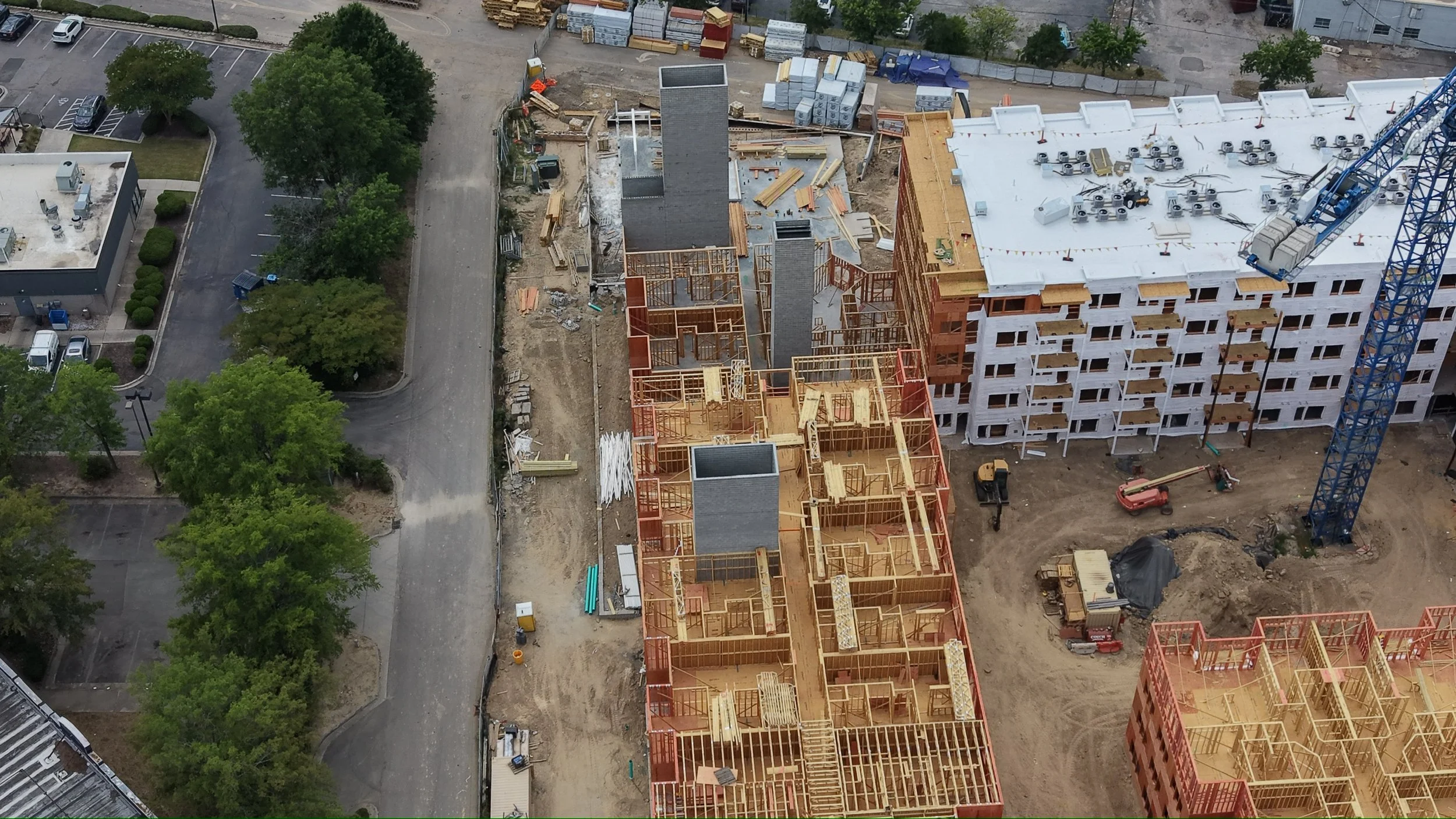 Aerial view of a construction site showing a partially built multi-story building, construction materials, and equipment with trees and parking lot nearby.