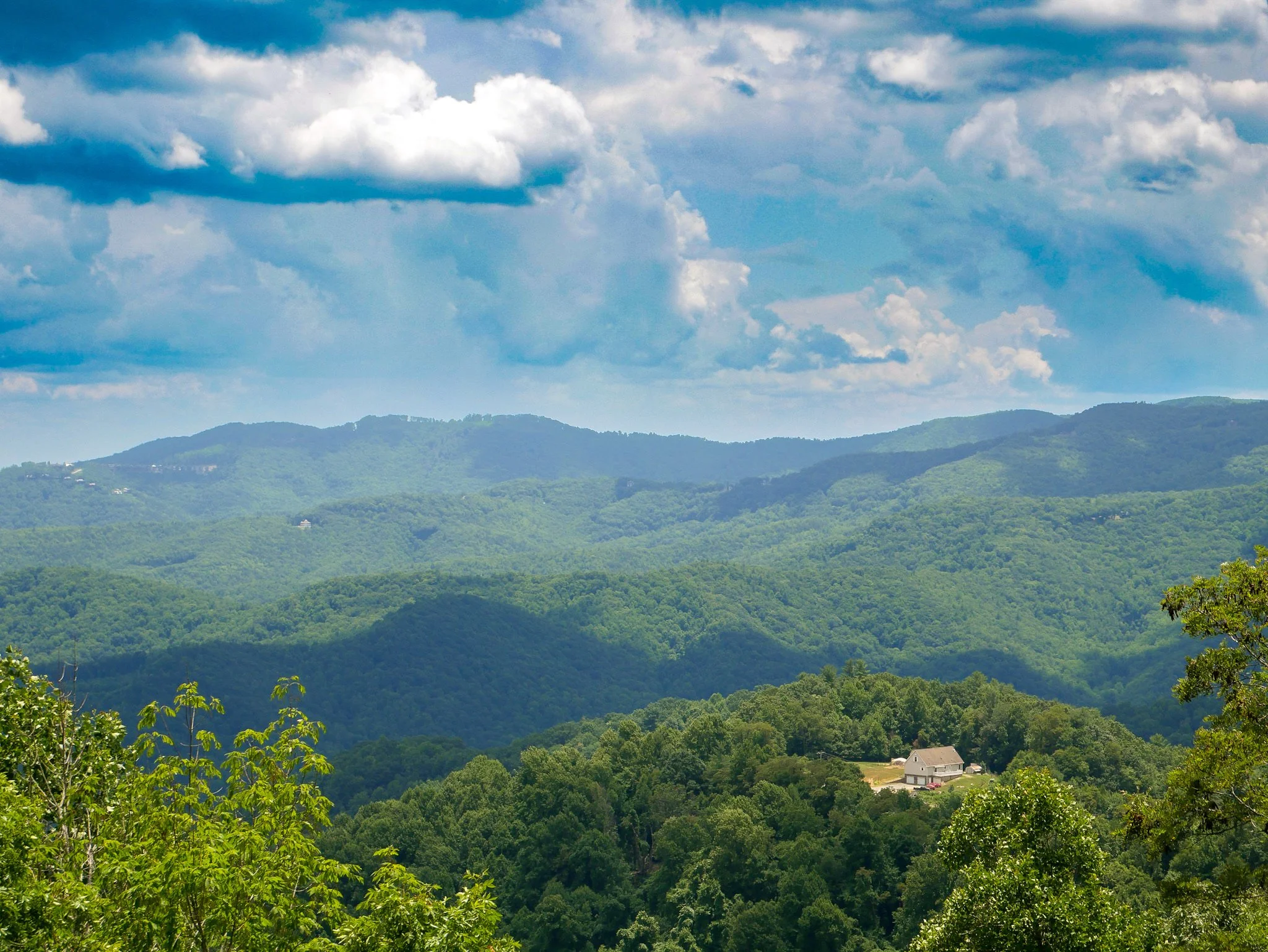 A scenic view of green rolling hills covered with trees, under a partly cloudy blue sky. A small house is visible near the bottom right.