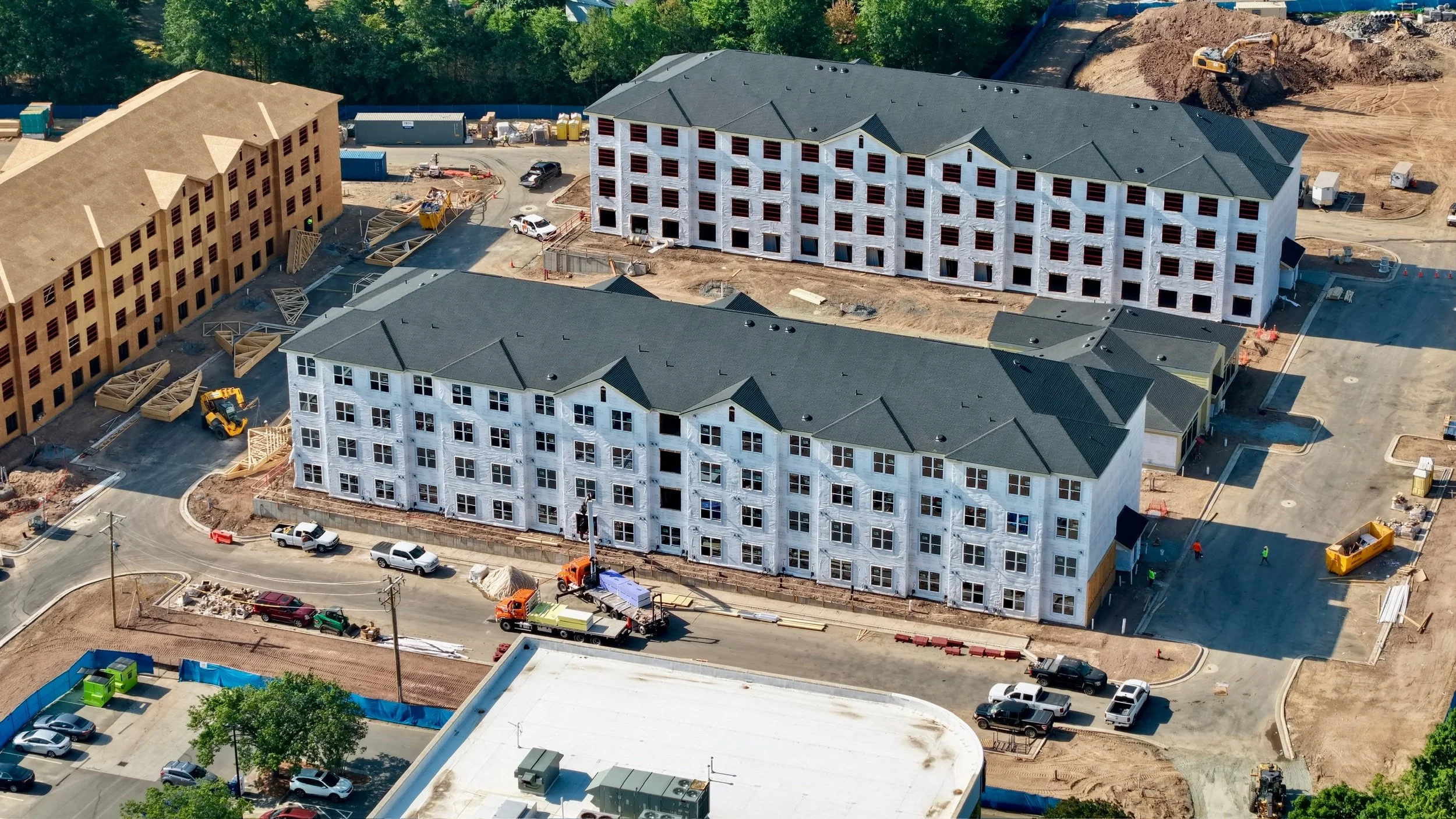 Aerial view of a construction site with multiple buildings under construction, construction vehicles, and surrounding parking lot with cars.