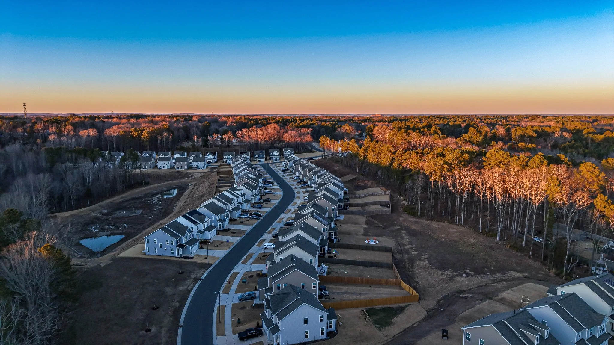 Aerial view of a suburban neighborhood with new white houses, a winding street, and a wooded area with trees in autumn colors under a clear blue sky during sunset.