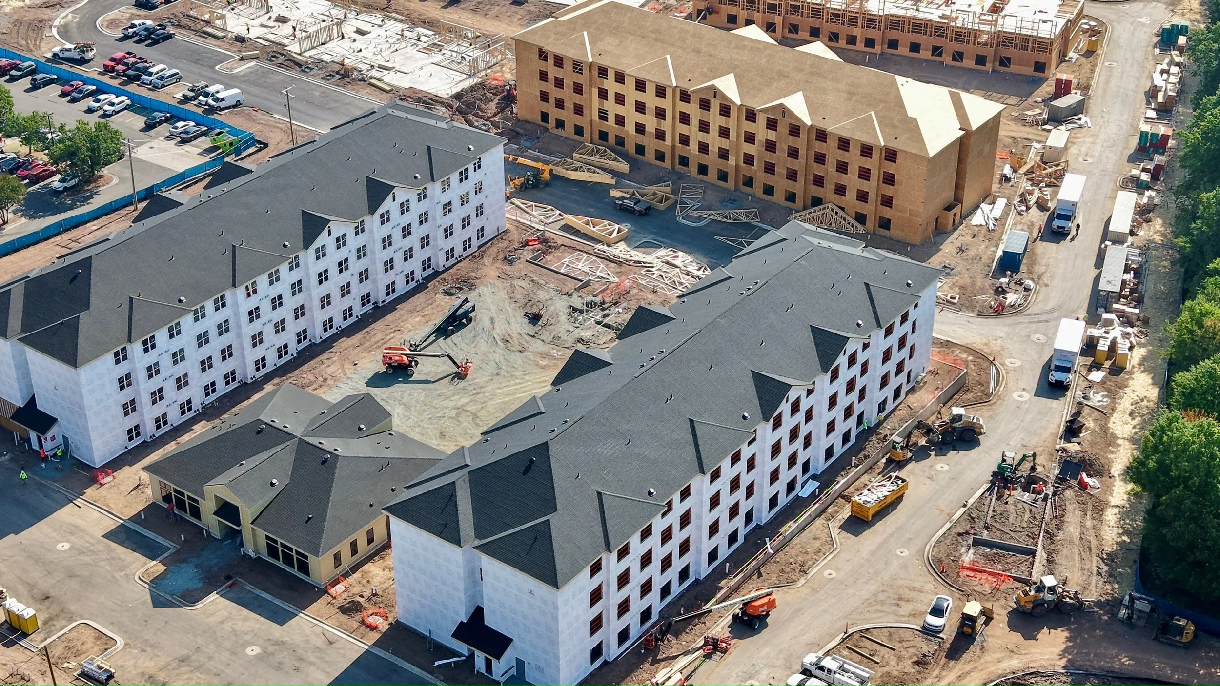 Aerial view of a construction site with multiple buildings in various stages of construction, several construction vehicles and equipment, parking lots, and surrounding roads.