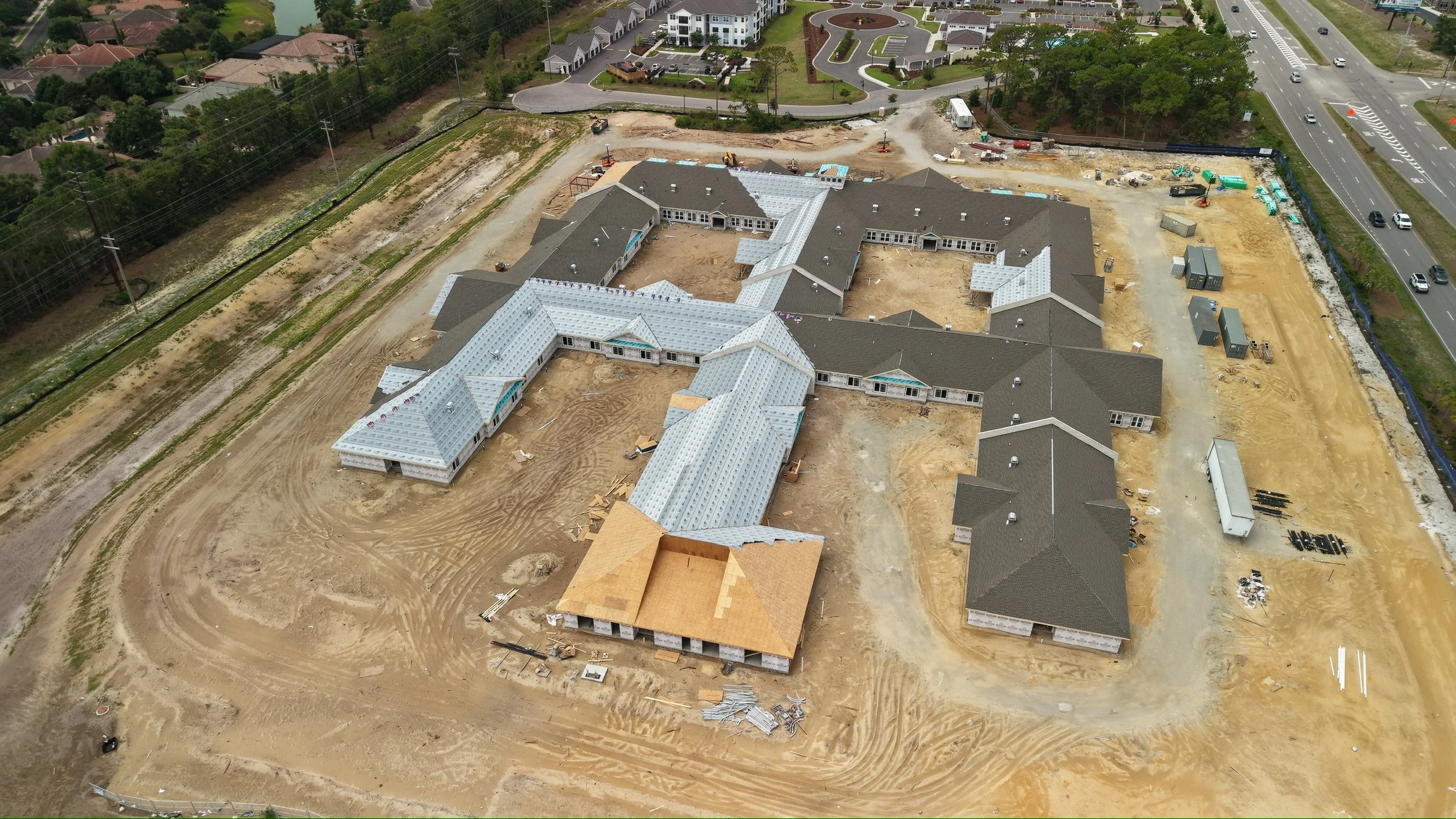 Aerial view of a construction site with several buildings in progress, surrounded by dirt and gravel, with completed roofs, some vehicles, and nearby residential area and roads.