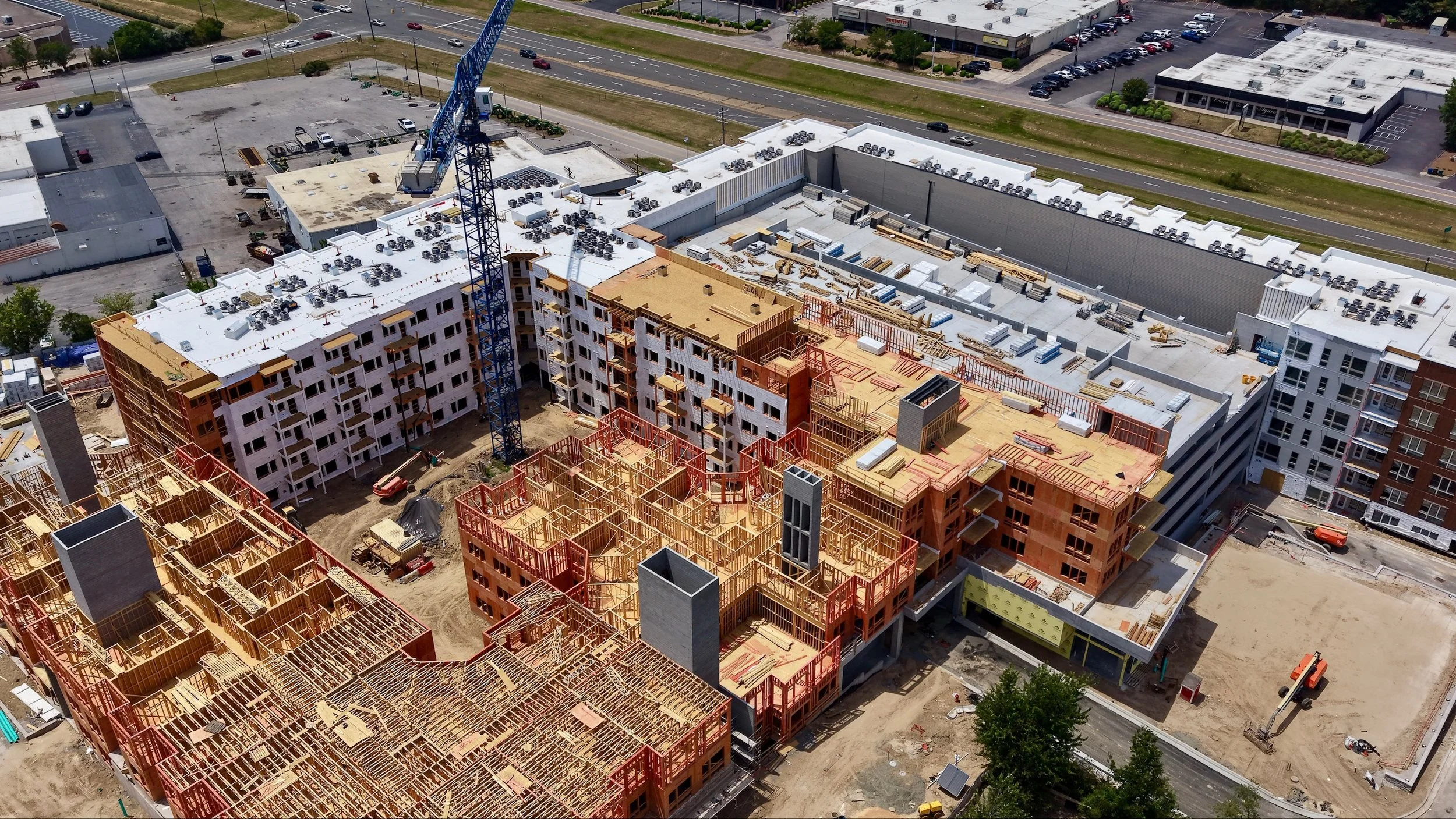 Aerial view of a construction site with multiple buildings in progress, including a high-rise with scaffolding and a crane, surrounded by roads and parking lots.