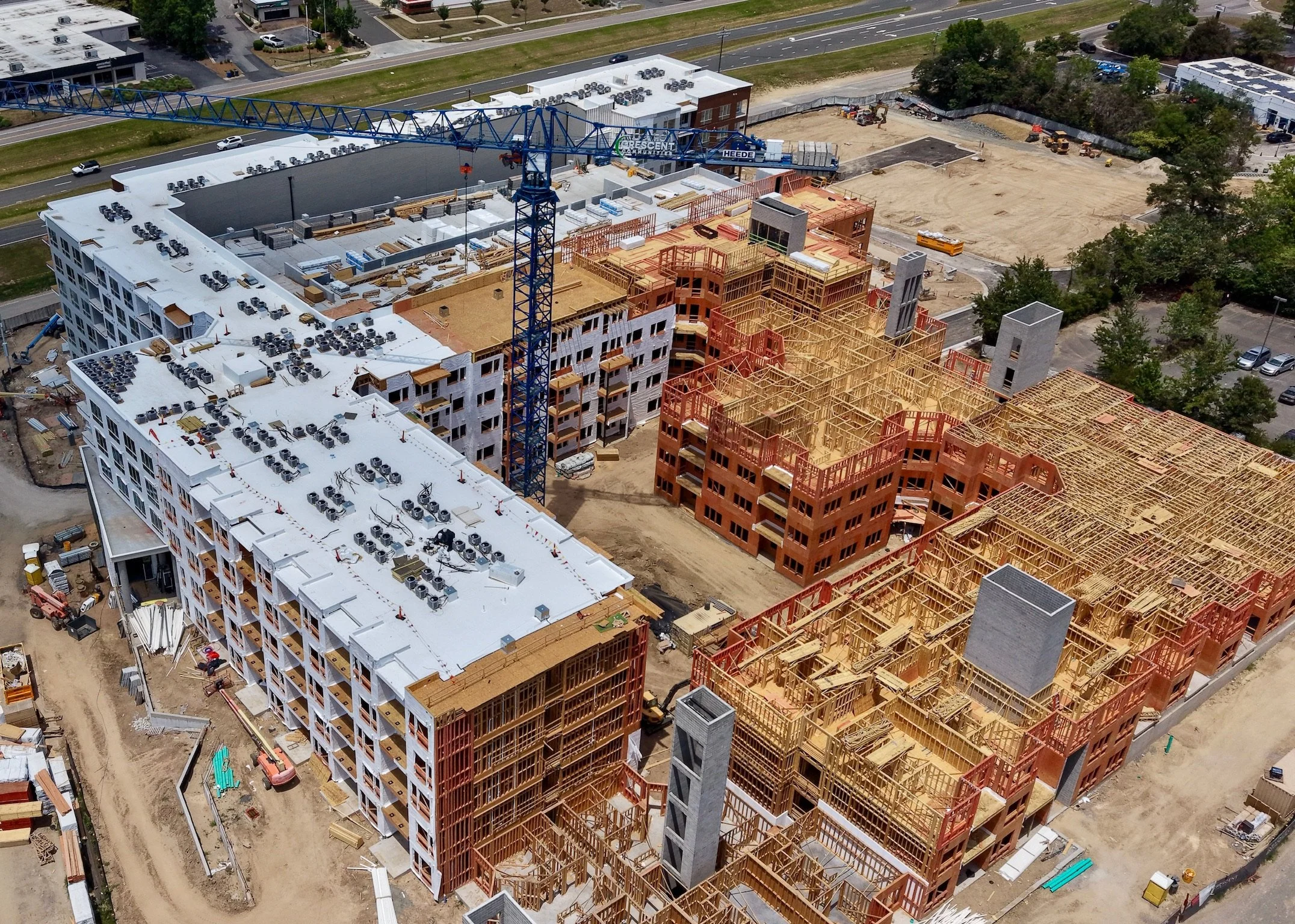 Aerial view of a large construction site where multiple buildings are under construction, with some having white roofing and others with exposed wooden frameworks, alongside a blue crane and construction equipment.