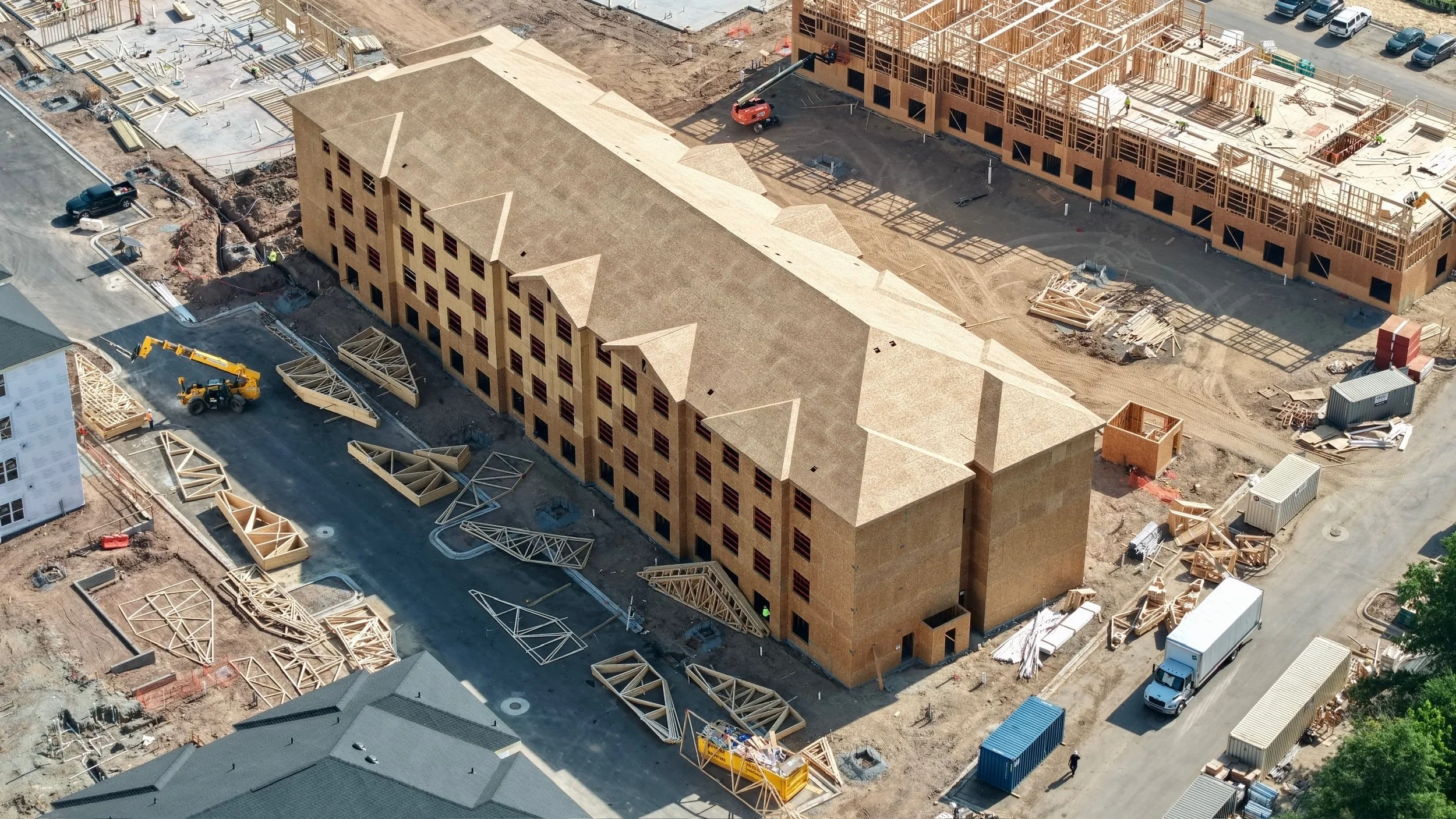 An aerial view of a construction site with multiple partially built brick buildings, construction materials, and workers. There are construction equipment and vehicles present, with some areas under active development.