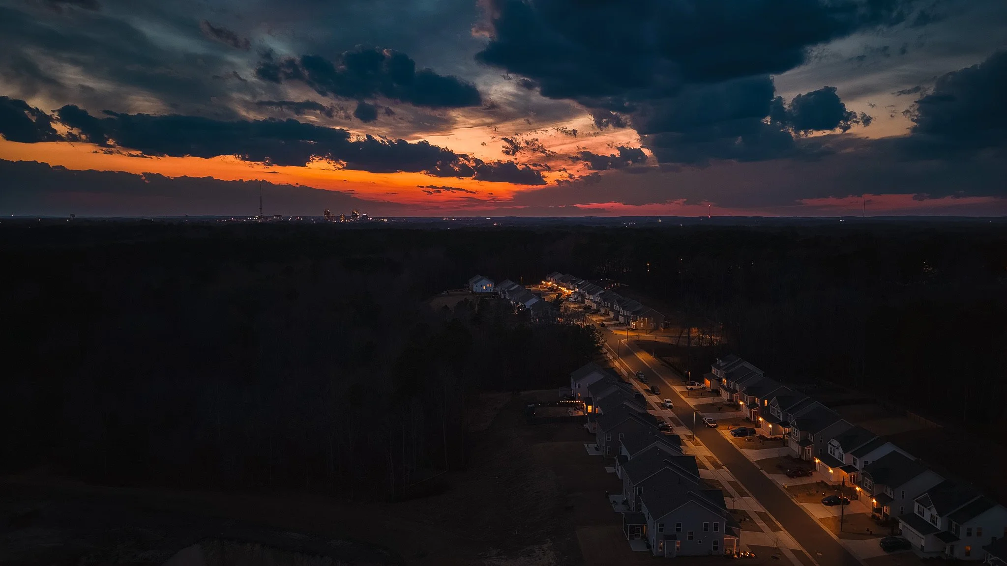 A sunset over a suburban neighborhood with houses and streets illuminated, dark clouds in the sky with orange and yellow hues at the horizon.