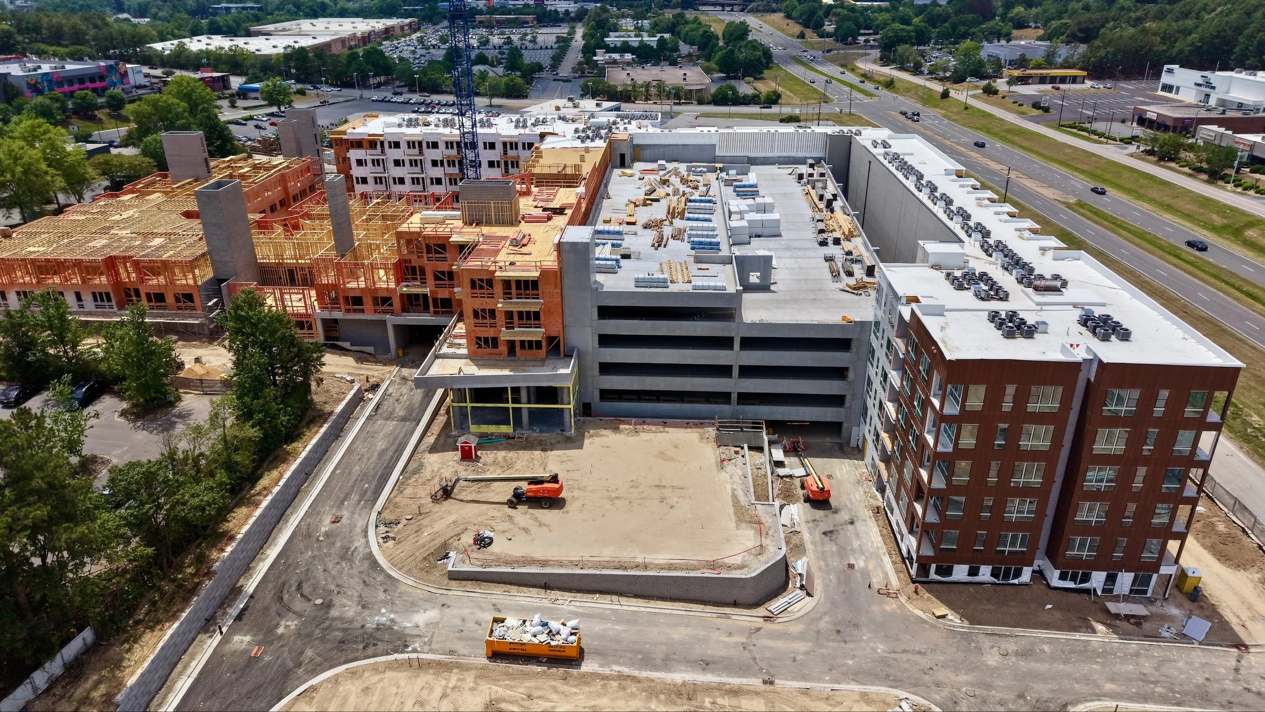 Aerial view of a multi-story building under construction with several cranes and construction equipment, surrounded by trees and parking lots.