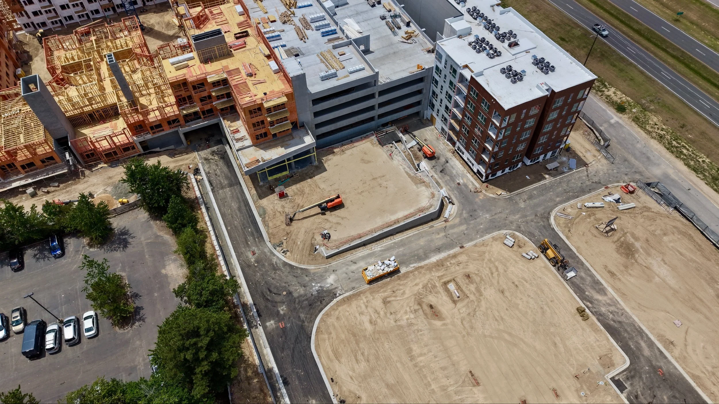 Aerial view of a construction site with several buildings in different stages of completion, trucks, and construction materials. There is a parking lot with cars on the left and a road on the right.