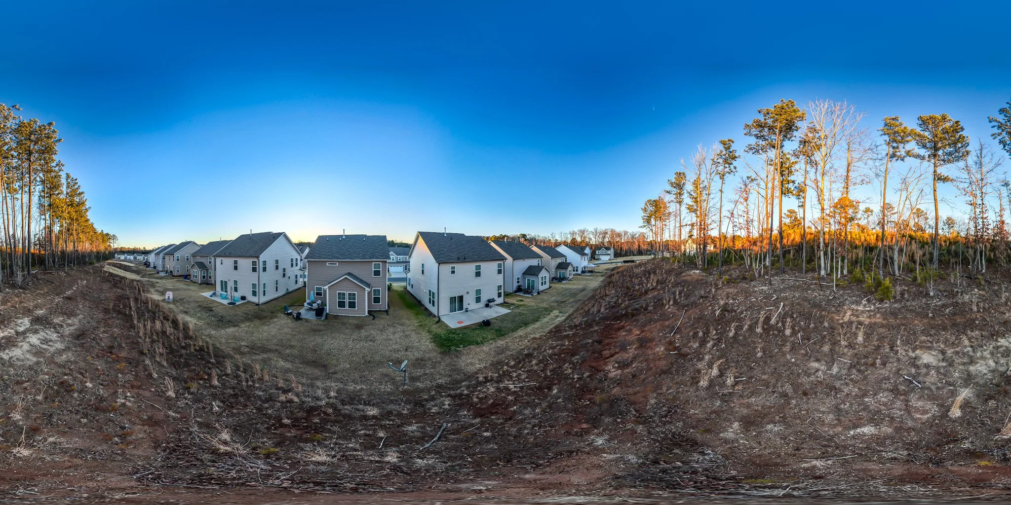 A panoramic view of a residential neighborhood with new houses, some with backyards, against a clear blue sky at sunset. There are trees on the right side and exposed soil in the foreground.