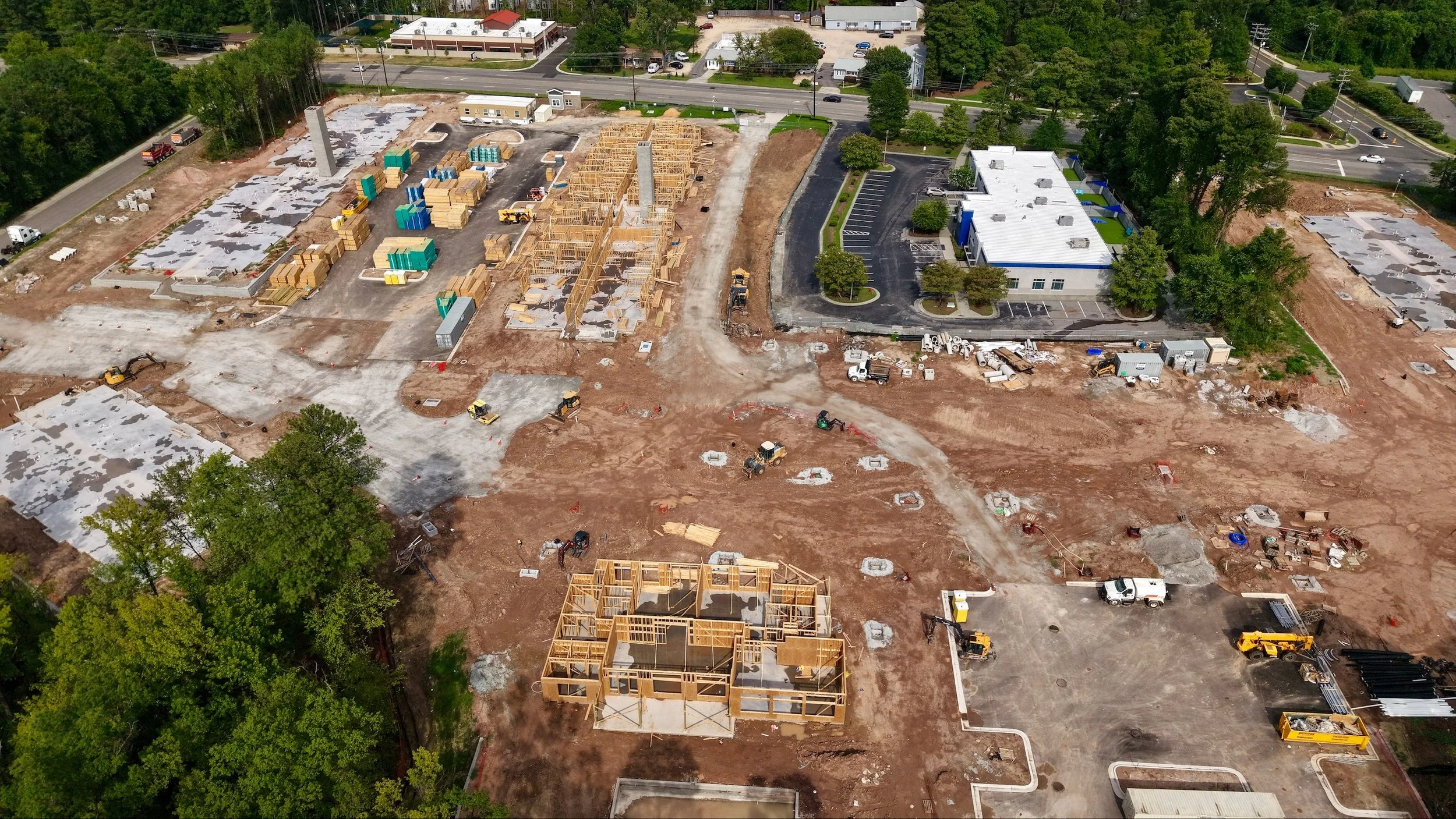 Aerial view of a construction site with wooden framing for buildings, construction machinery, and materials, surrounded by trees and nearby roads.