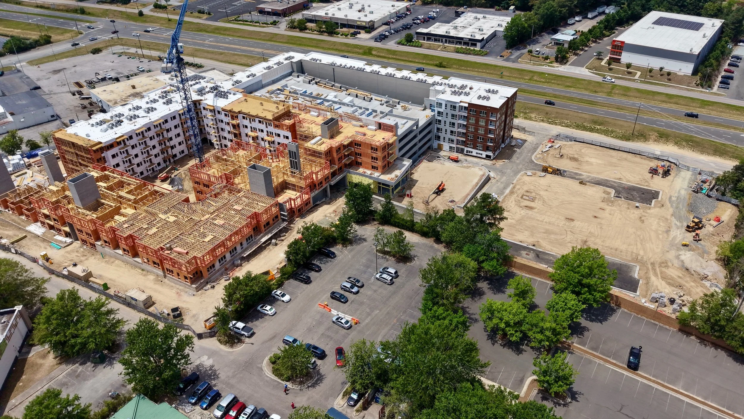An aerial view of a construction site with buildings under construction, a crane, and surrounded by parking lots and roads.