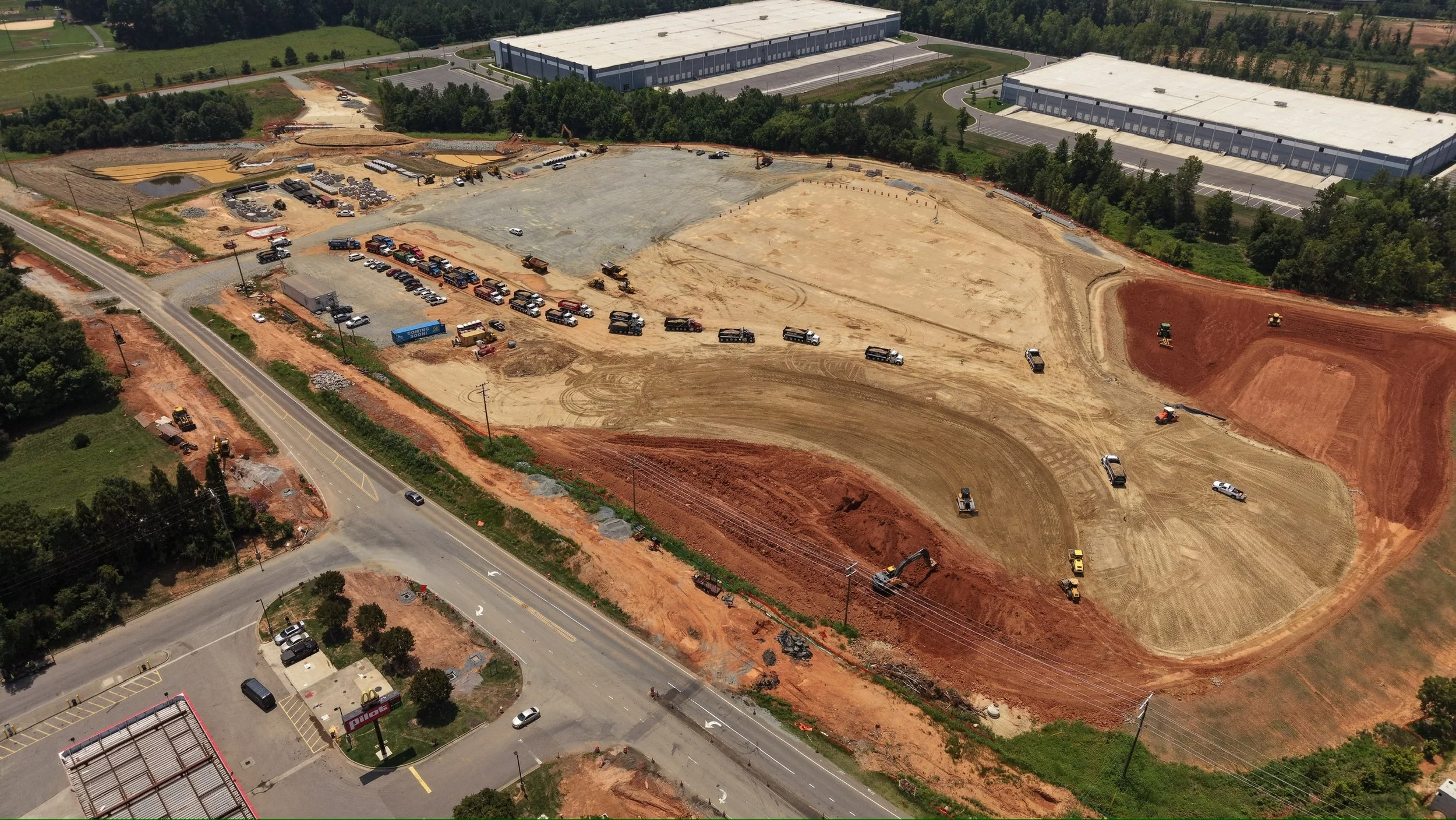 Aerial view of a construction site with multiple dirt areas, construction vehicles, and equipment, with a large warehouse building and parking lot in the background.