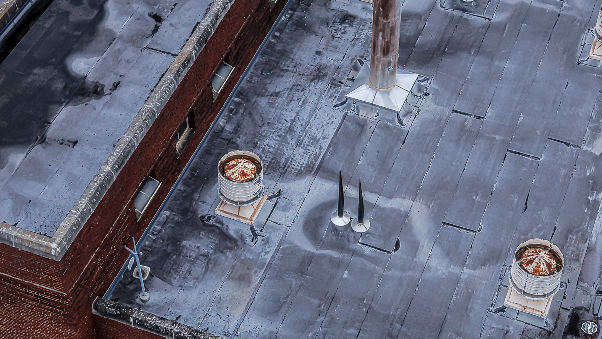 Aerial view of a building rooftop with black shingles, brick walls, two cylindrical exhaust vents, a central vent, a chimney, and two cleats.