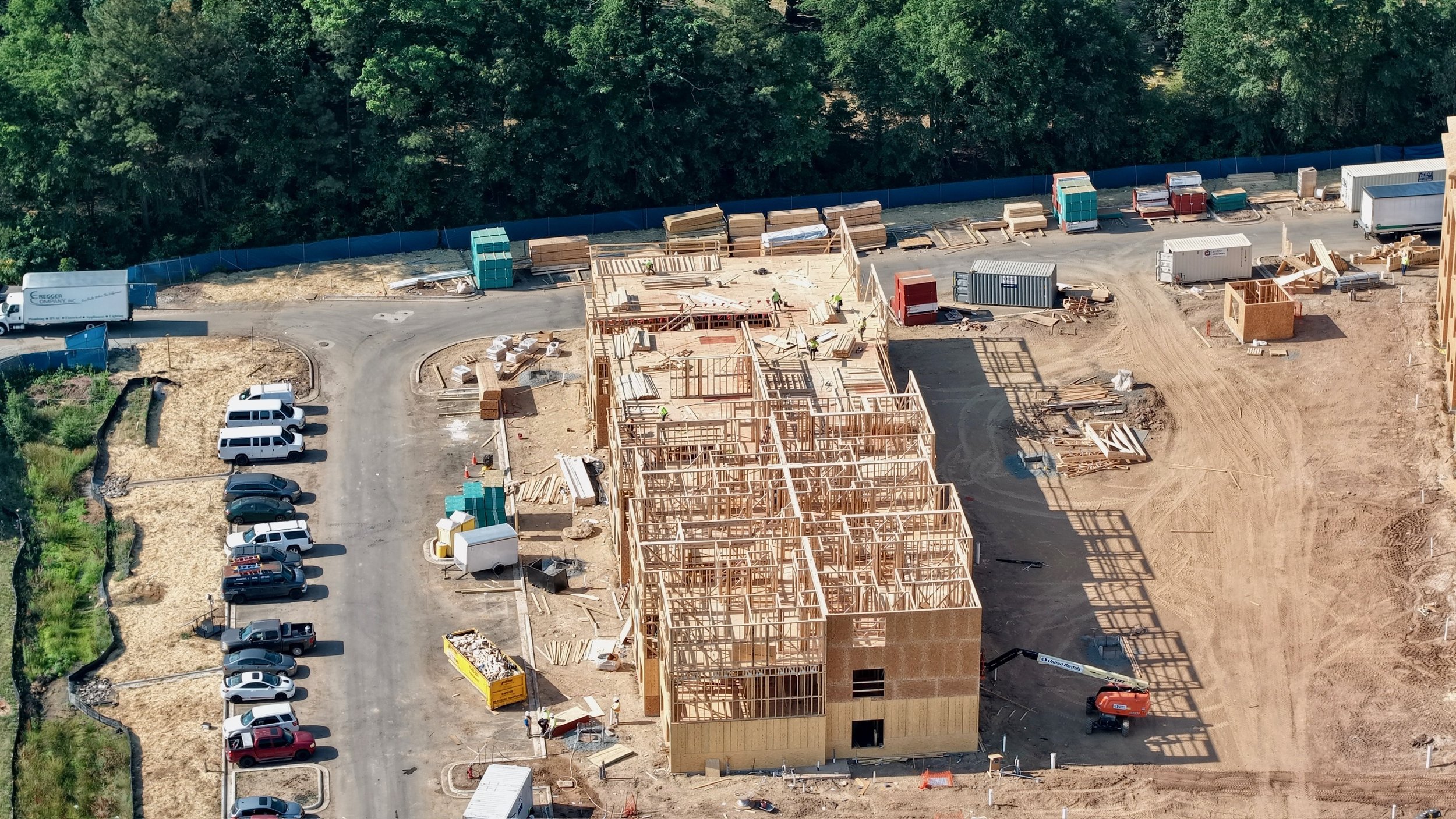 An aerial view of a construction site with wooden framing for a building, construction workers, construction materials, parked cars, trucks, and containers, with trees lining the background.
