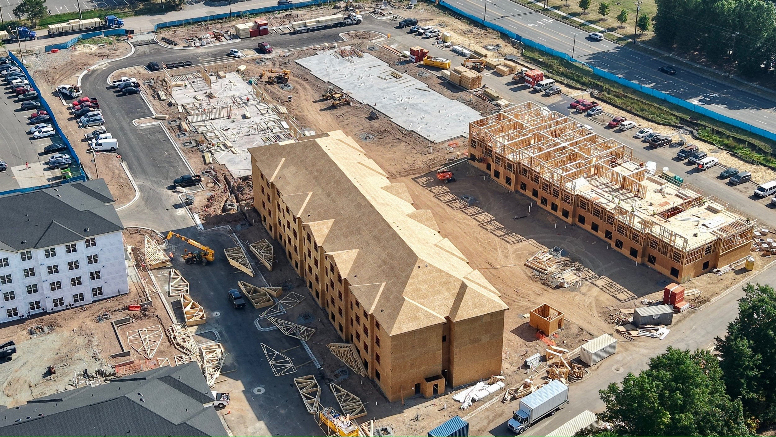 Aerial view of a construction site with partially built wooden structures, construction vehicles, and parked cars, near a parking lot and a road.