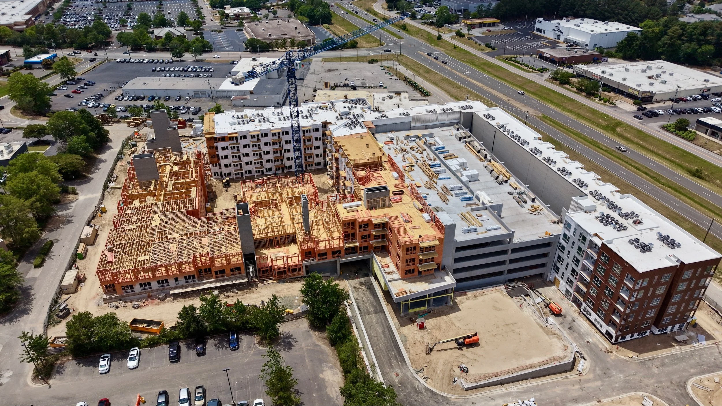 Aerial view of a construction site with partially built multi-story buildings, a crane, and surrounding parking lots and roads with sparse traffic.