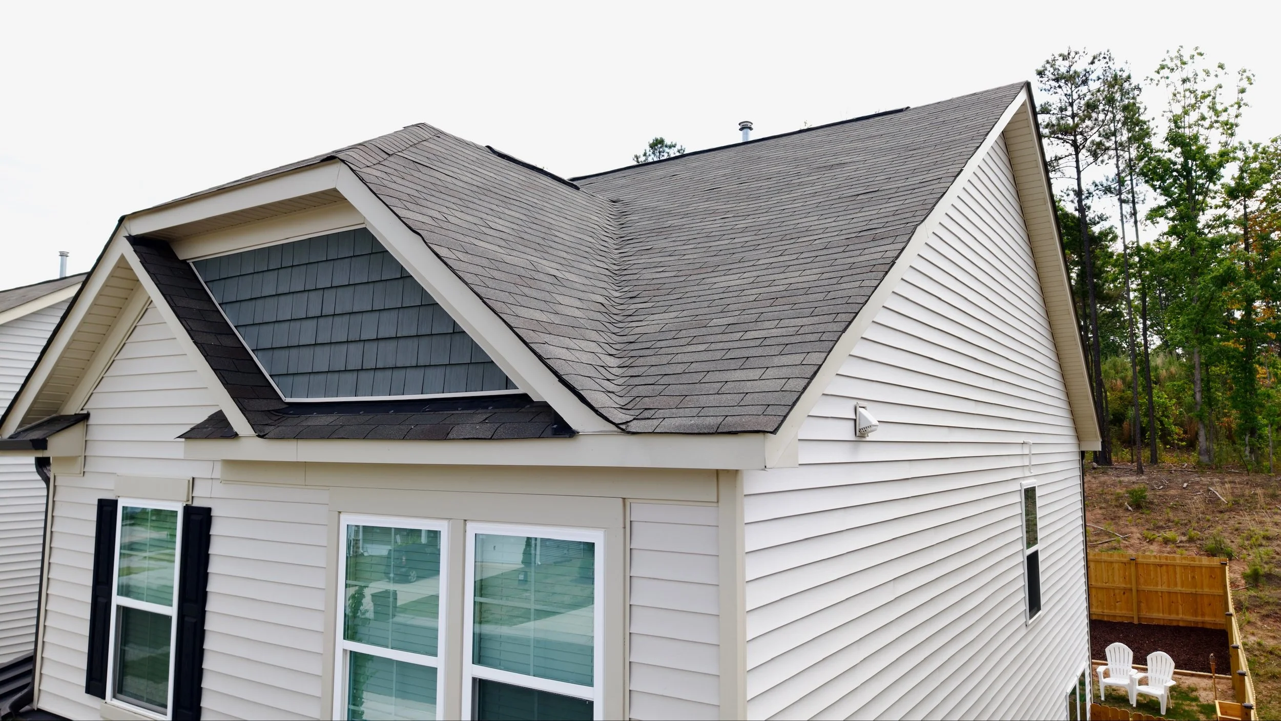 A house with white siding, black shutters, and a steep gray shingle roof. A window with multiple panes and a black shutter is visible on the lower left. There are trees and a wooden fence in the background.