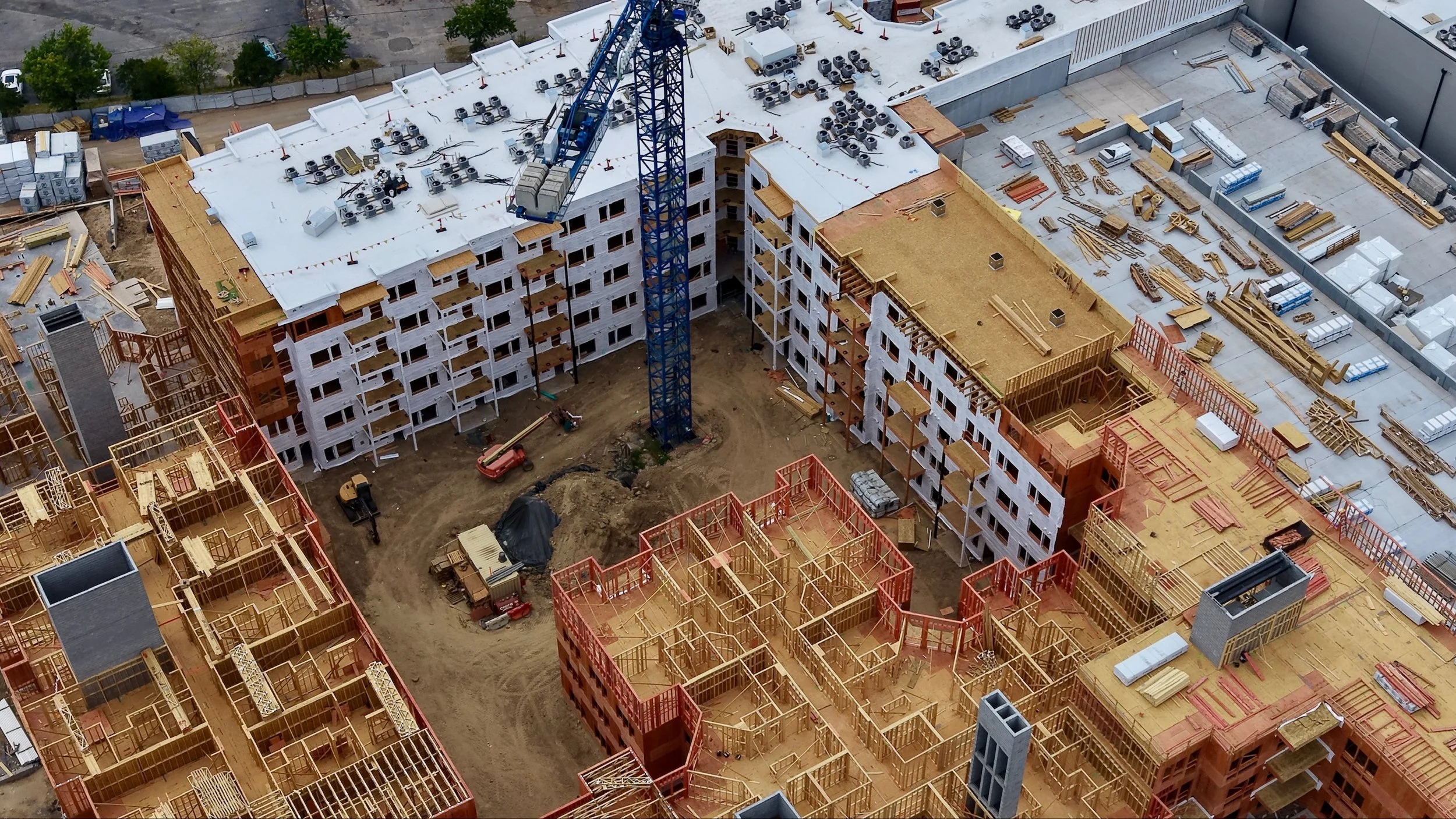 Aerial view of a cluttered construction site with partially built multi-story buildings, construction materials, and equipment including a blue tower crane, wood framing, and concrete blocks.
