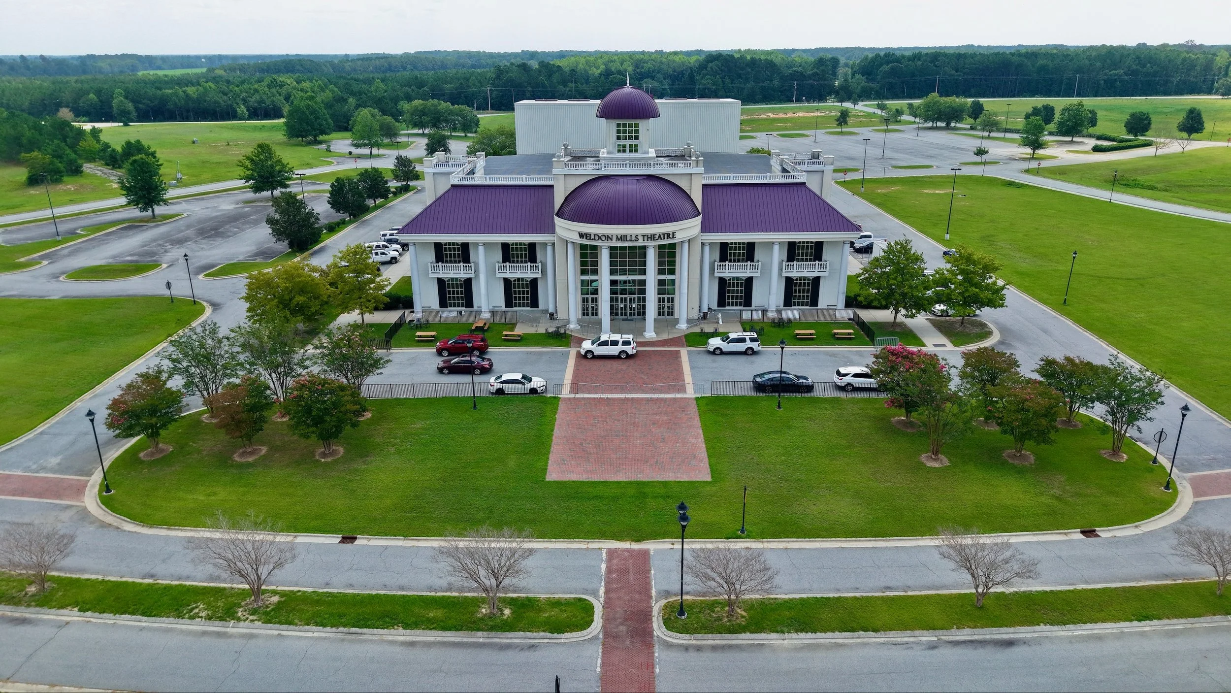 Aerial view of a large white building with purple roofs and columns, labeled Weldon Mills Theatre. The building is surrounded by green lawns, trees, parking lots, and empty streets, with a forest and open fields in the background.