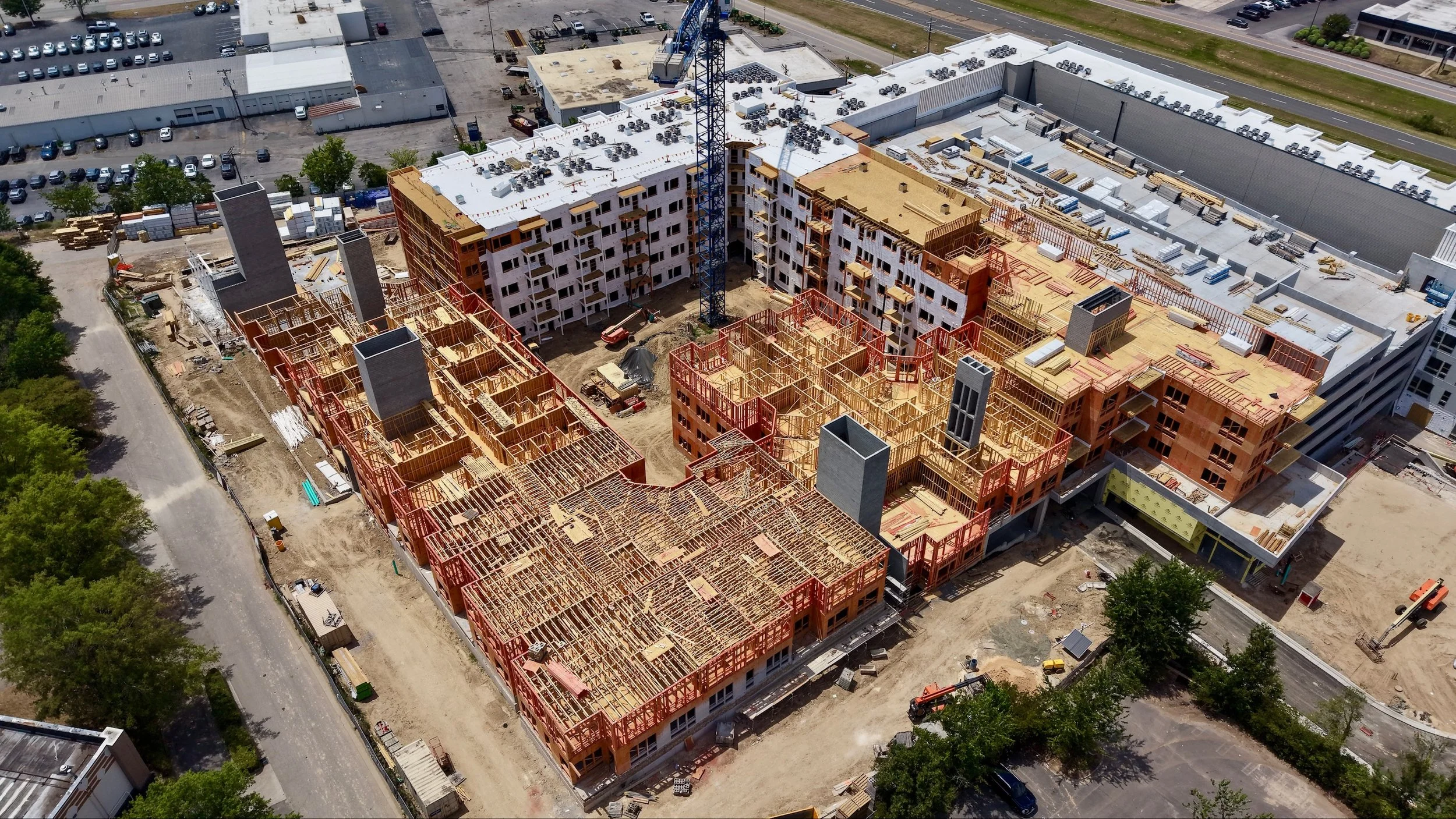 An aerial view of a construction site with multiple partially built multi-story buildings, construction materials, and a crane, surrounded by parking lots and roads.