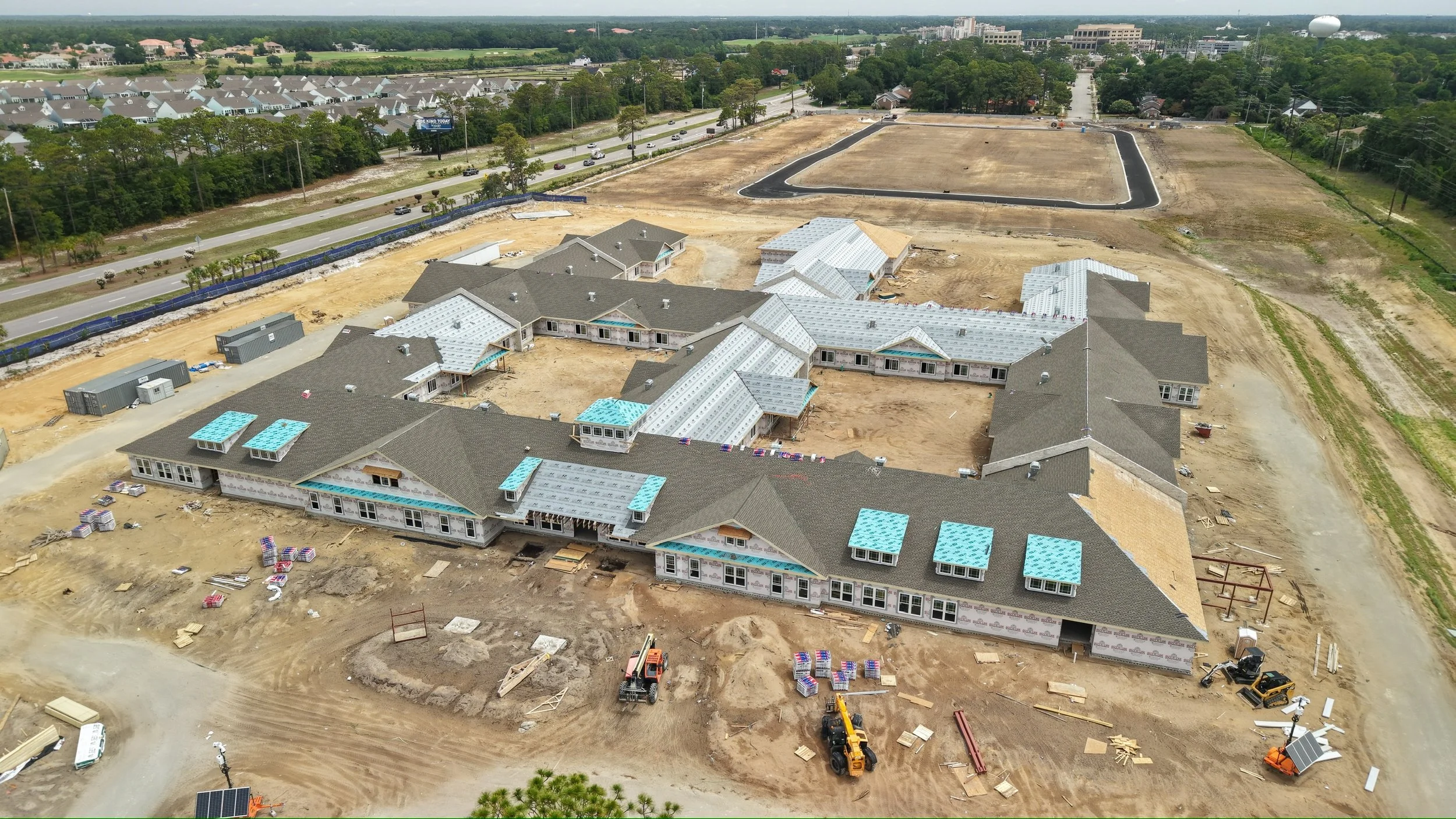 Aerial view of residential house construction site with partially completed buildings, construction machinery, and surrounding roads and green areas.