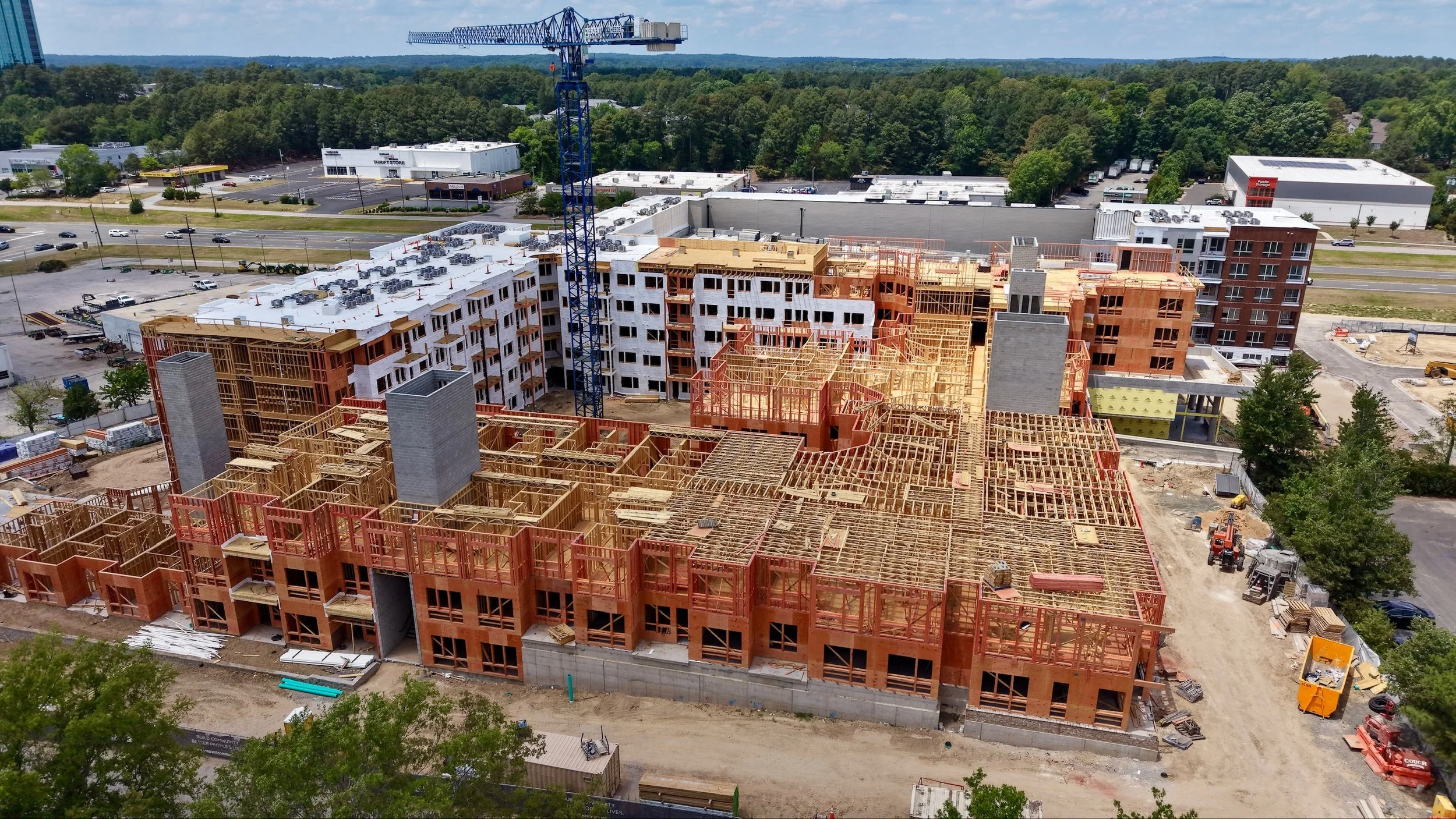 Aerial view of a large construction site with multiple buildings under construction, showing wooden framing and some completed walls, surrounded by trees and highways in the background.
