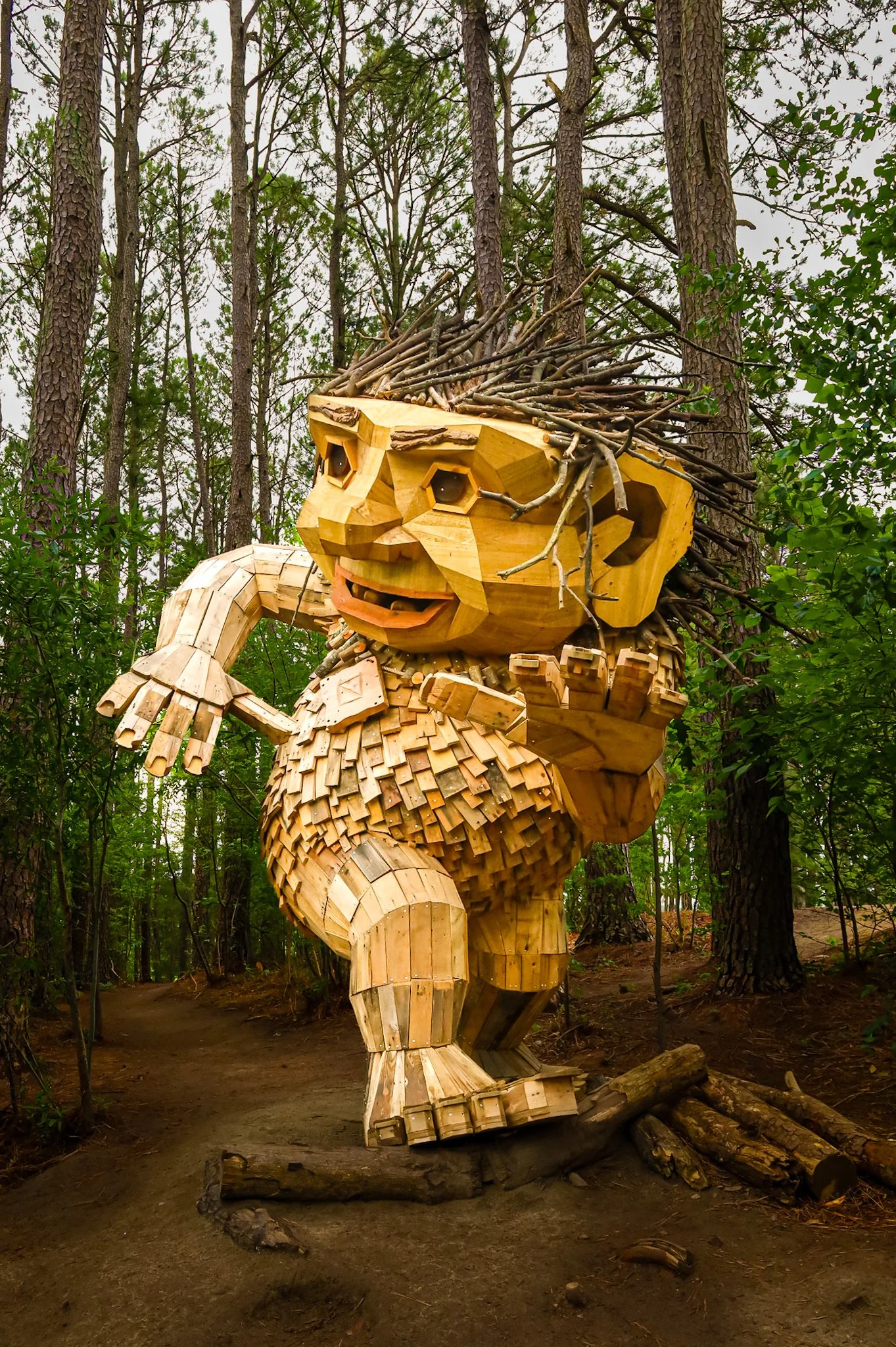 Wooden sculpture of a lion in a forest, with a mane made of sticks and branches, and the lion has a fierce expression.