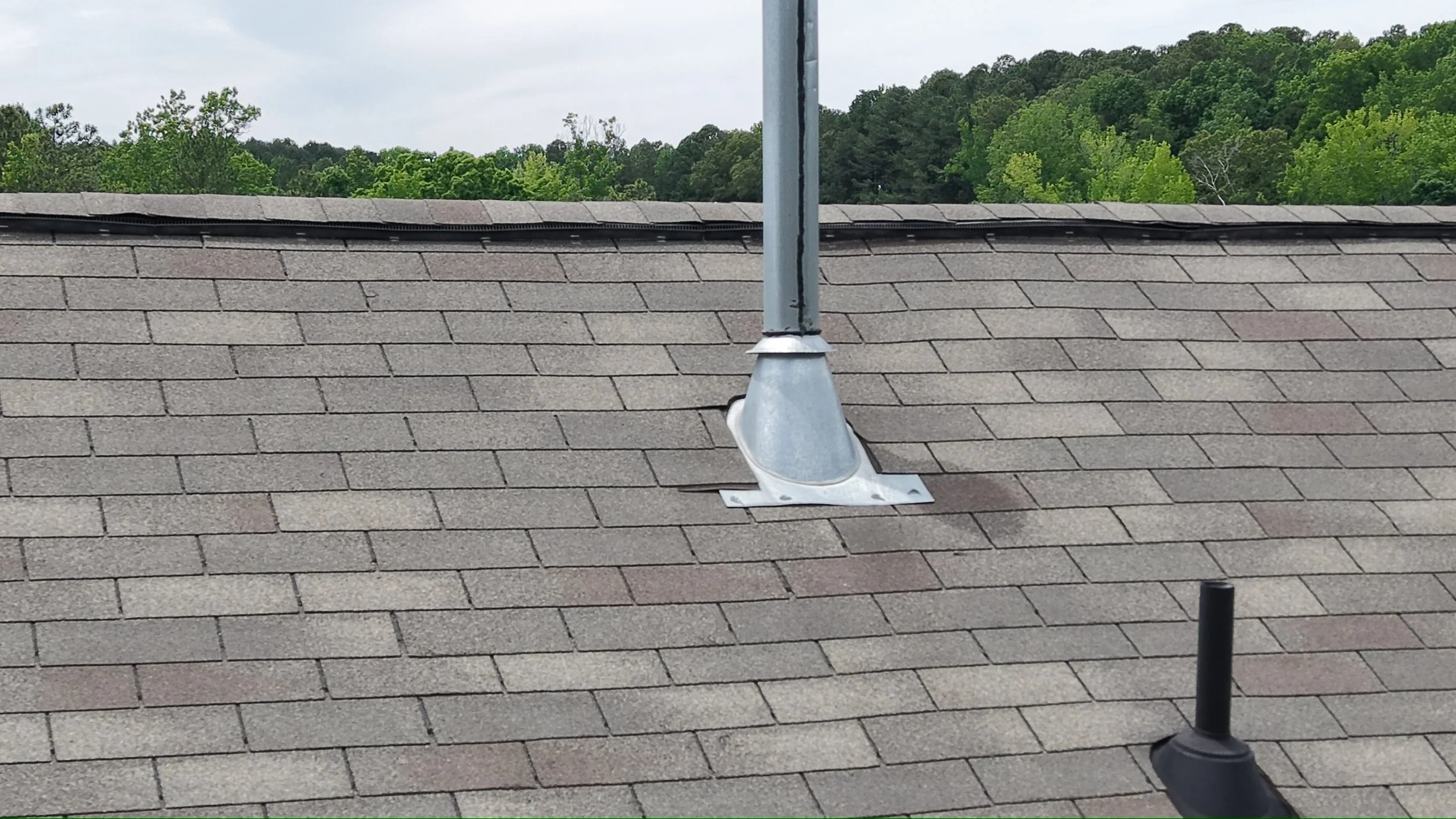 A rooftop with asphalt shingles, a metal vent pipe with a flashing base, and a weather vane post against a background of green trees and a cloudy sky.