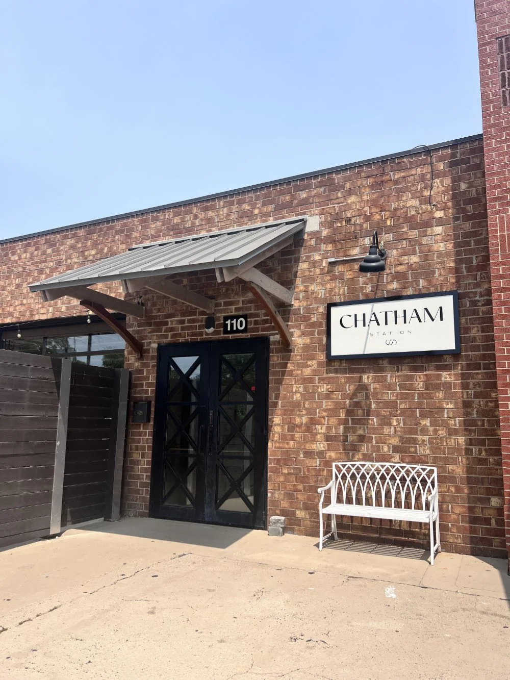 Brick building entrance with black double doors, wood awning, and white bench next to a sign reading 'Chatham Station US', under a clear blue sky.