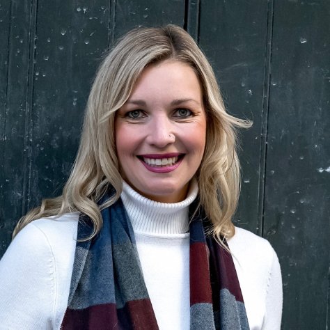 A woman with long blonde hair, smiling, wearing a white turtleneck, and a multicolored scarf, standing in front of a dark textured wall.